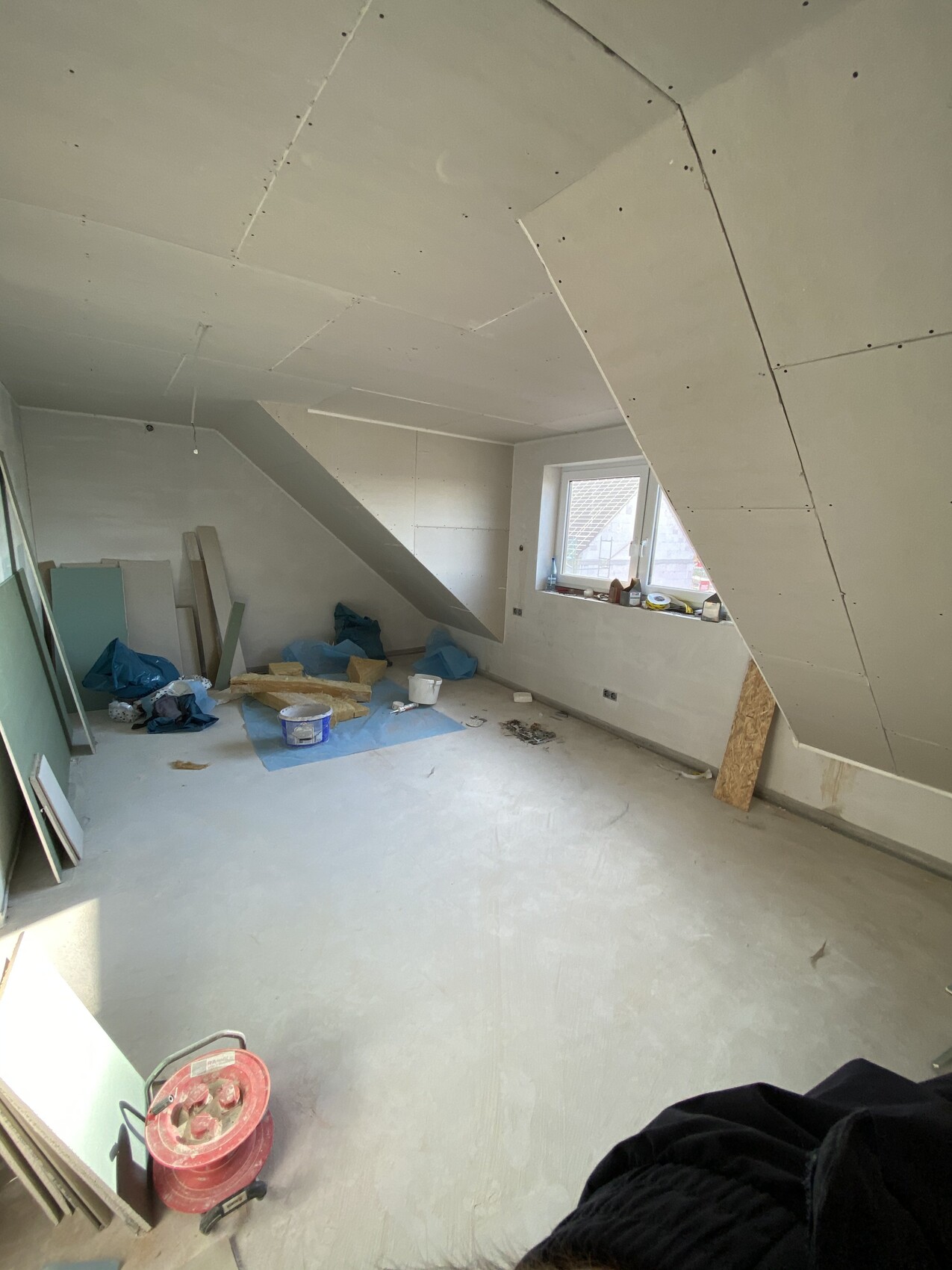 Unfertiger Dachbodenraum mit Trockenbauwänden, Baumaterialien, Werkzeugen und Fenster.