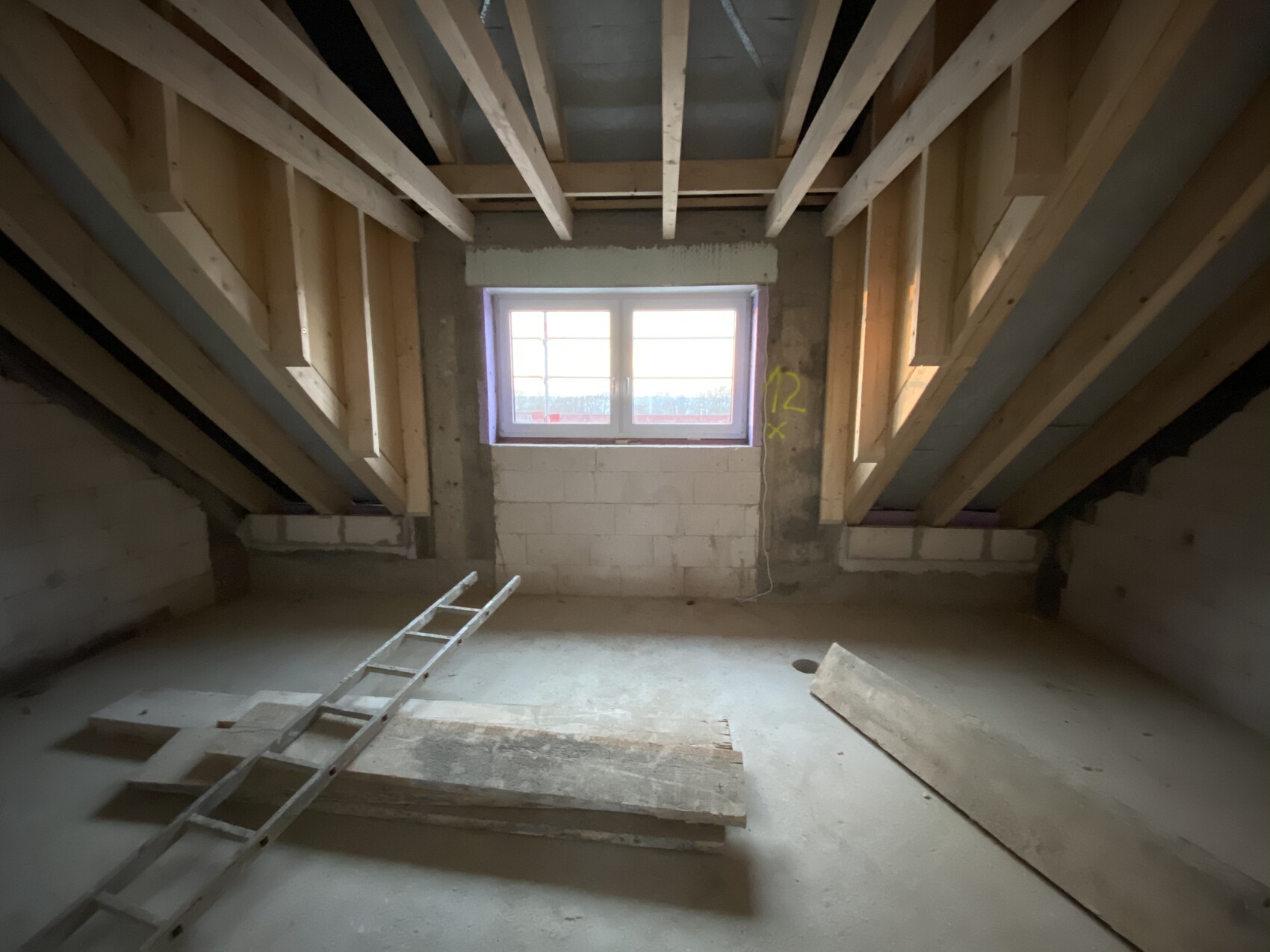 Dachboden im Rohbau: Holzbalken, Fenster in der Mitte, Leiter und Baumaterialien auf dem Boden.
