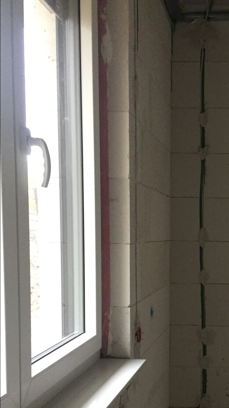 Weißer Fensterrahmen mit Griff; unfertiger Innenraum aus Betonblöcken und Rohrleitungen sichtbar.