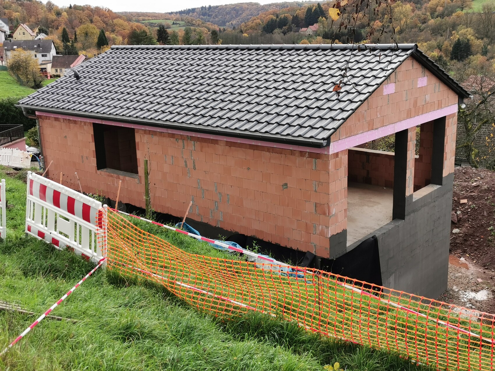 Rohbau eines Hauses auf Hang mit grauem Dach, Bauabsperrungen und Herbstlandschaft.