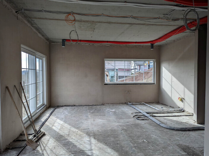 Baustellen-Innenraum: roher Boden, sichtbare Leitungen und rotes Rohr, Fenster, Schaufel an der Wand.