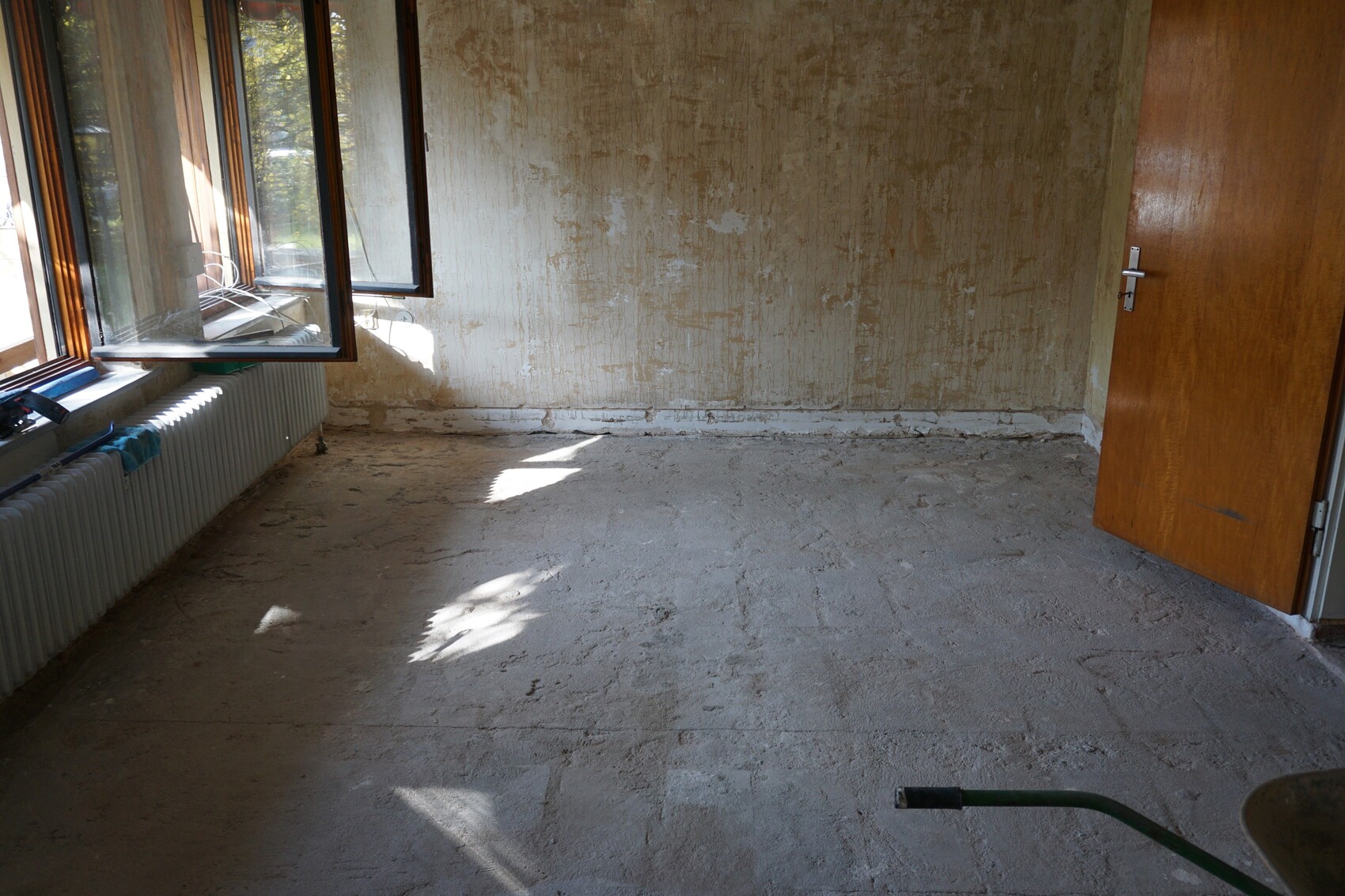 Renovierungsraum: leeres Zimmer, offene Fenster, rauhe Wände, Heizkörper, Holztür.