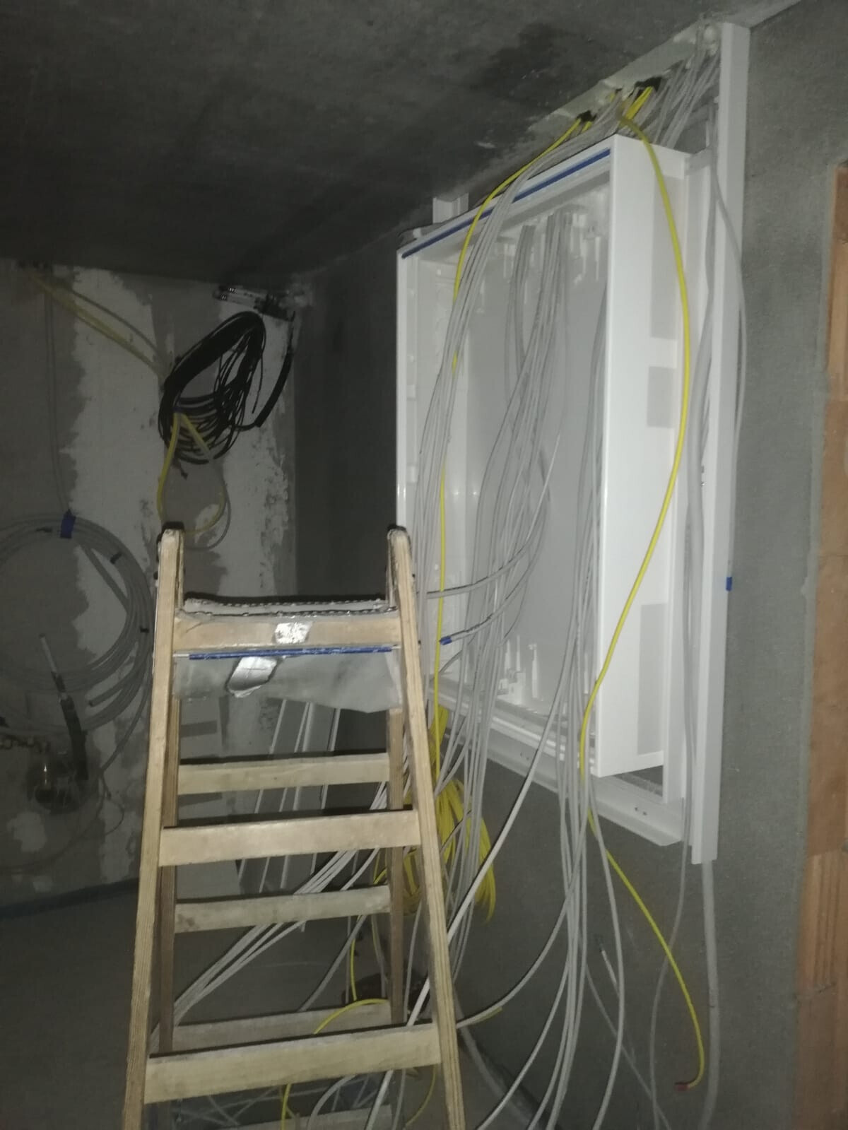 Verteilerkasten an der Wand mit vielen gelben und grauen Kabeln; Holzleiter davor.