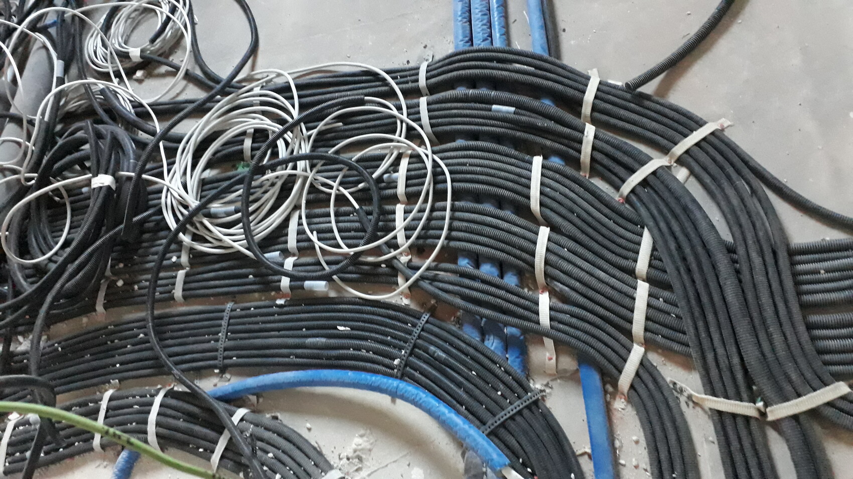 Viele schwarze Kabelbündel und lose Kabel auf dem Boden, dazu blaue Rohre und weiße Gurtbänder.