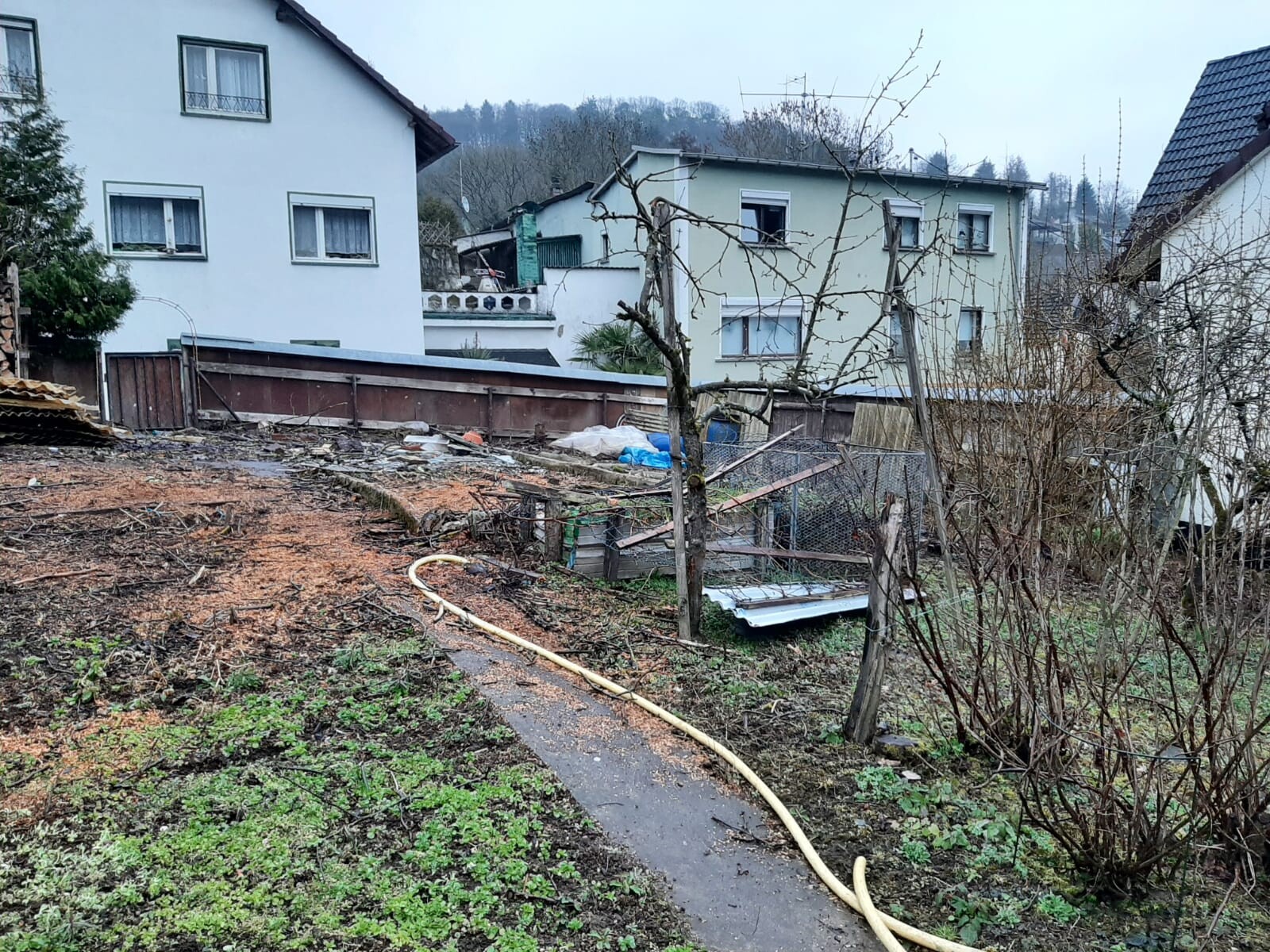 Garten mit kahlen Bäumen, langer Schlauch, Müll und Holzreste; Häuser im Hintergrund.