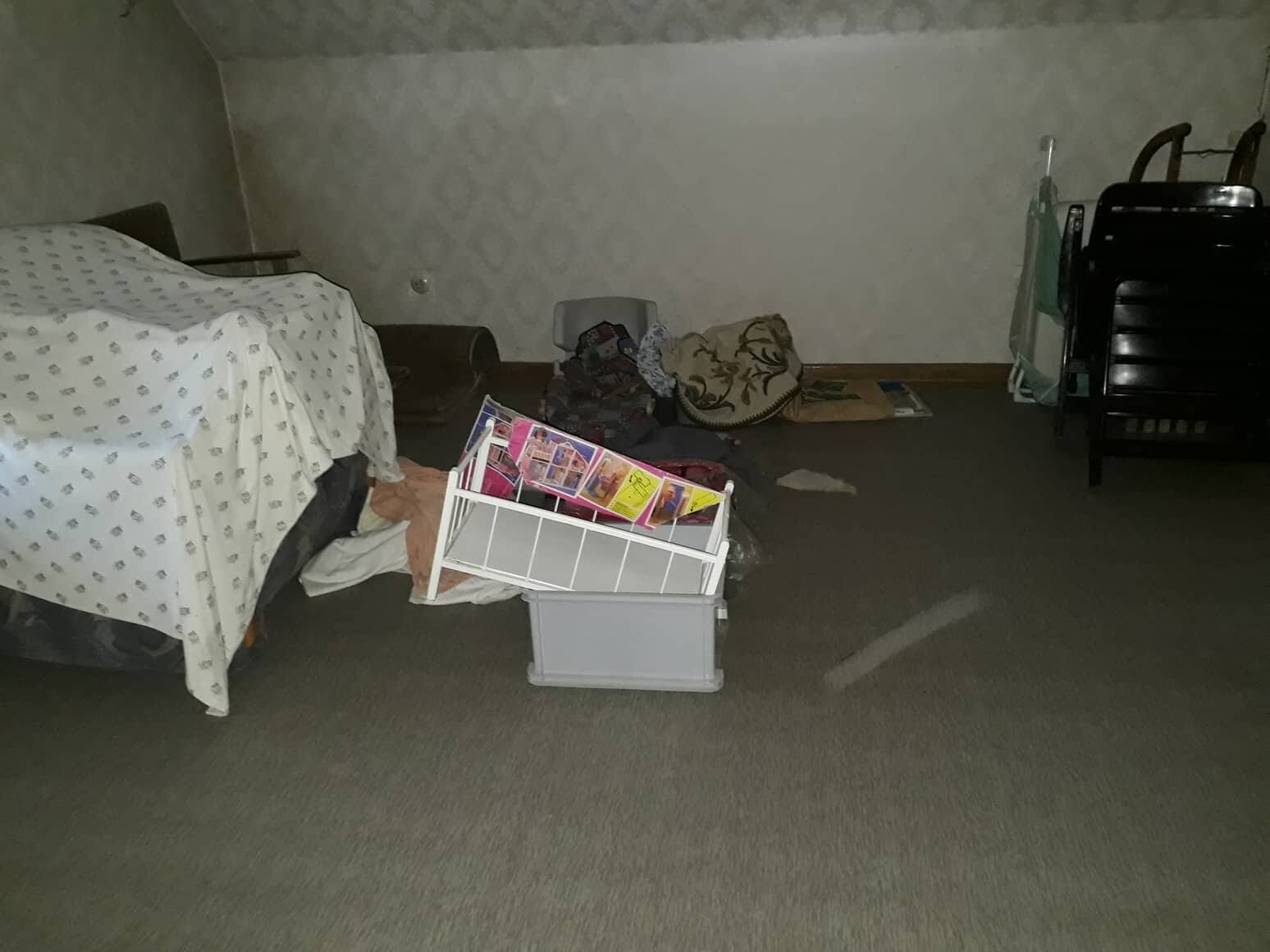 Unordentlicher Dachboden/Keller mit Bett, Decken, Kisten und einem umgekippten Aufbewahrungsregal.
