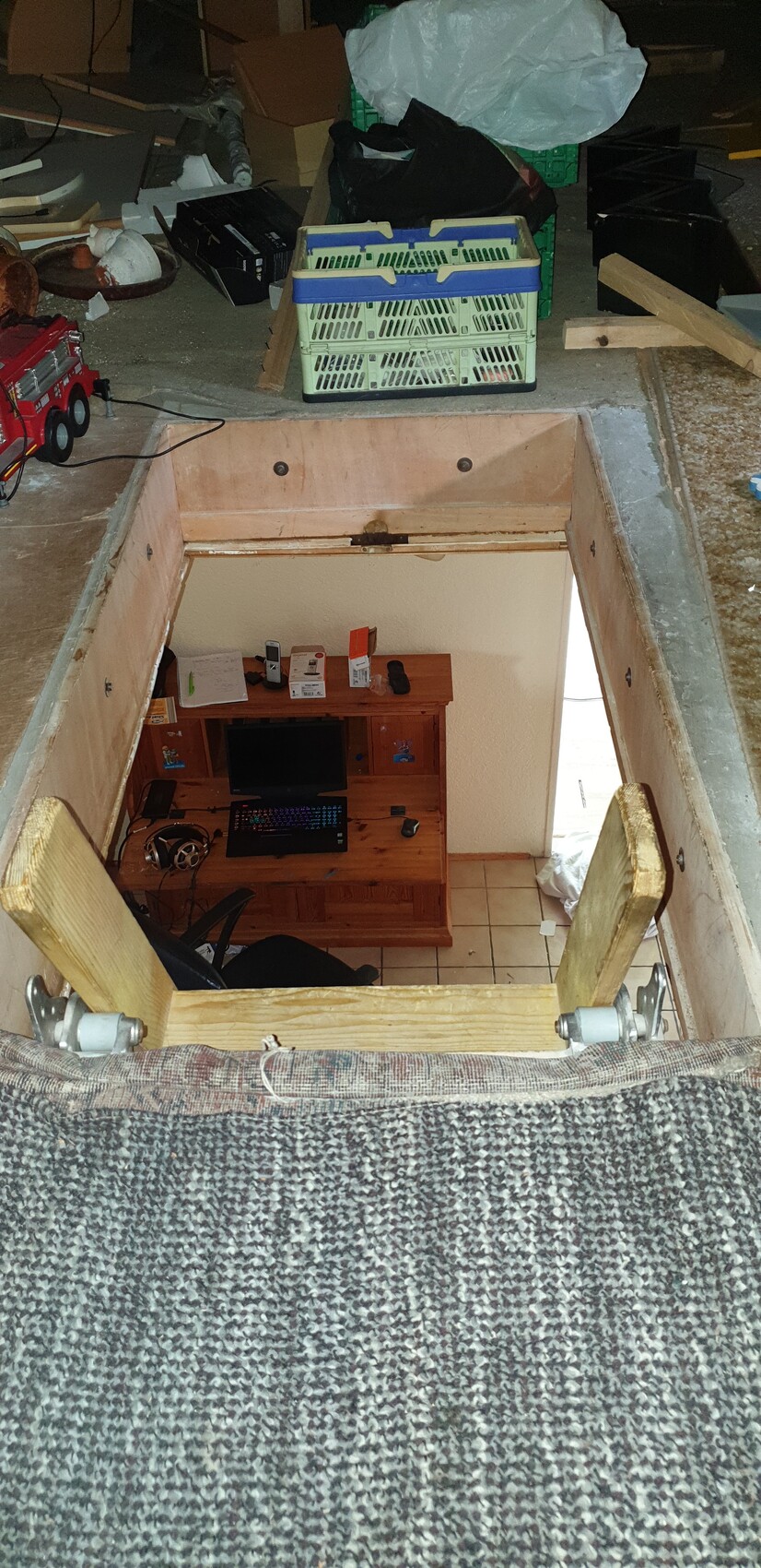 Offene Bodenluke führt in einen Kellerraum mit Schreibtisch, Computer, Monitor, Tastatur; Chaos oben.