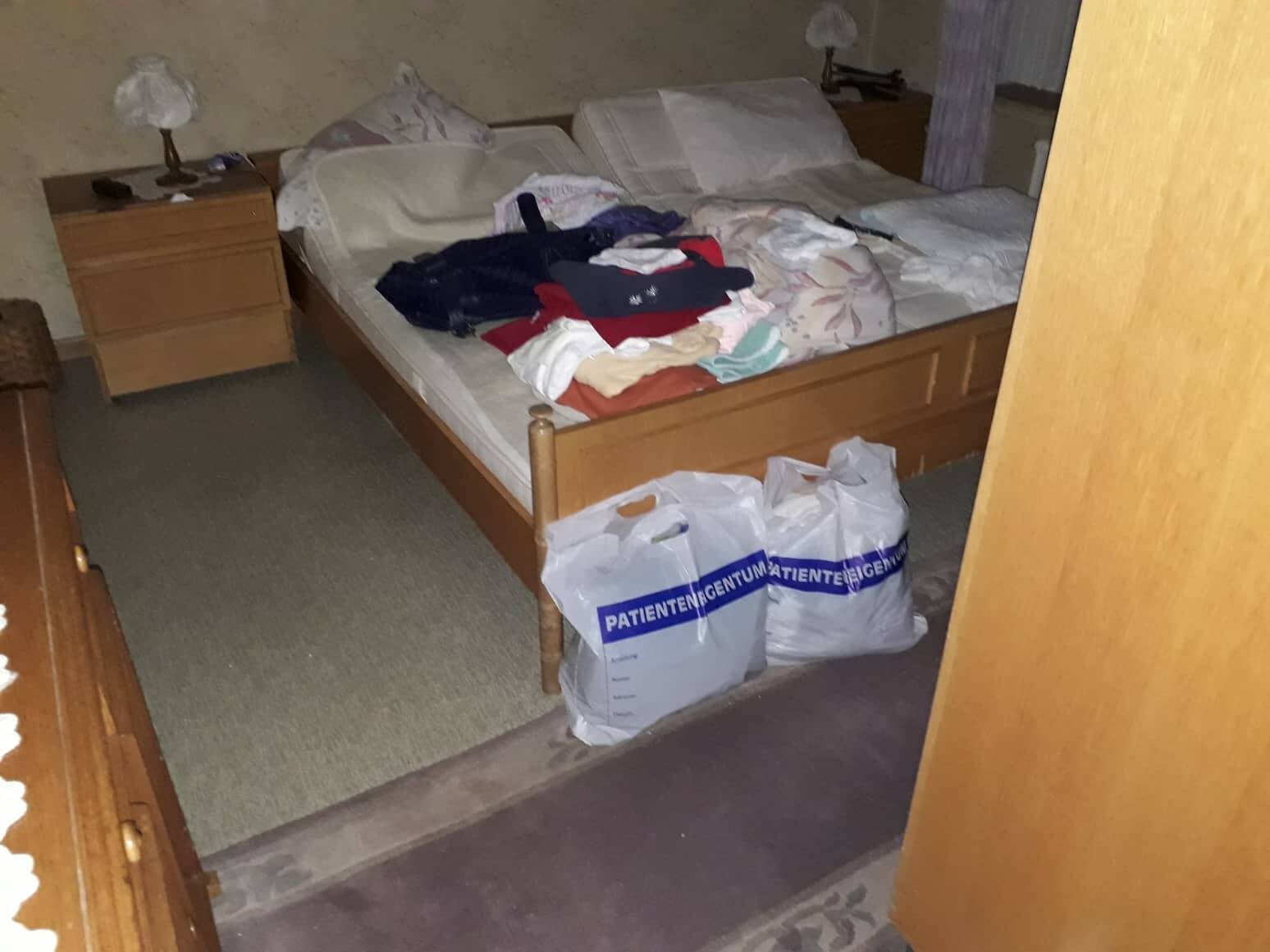 Unordentliches Schlafzimmer: Bett mit Kleiderstapel, zwei Plastiktüten am Fußende.