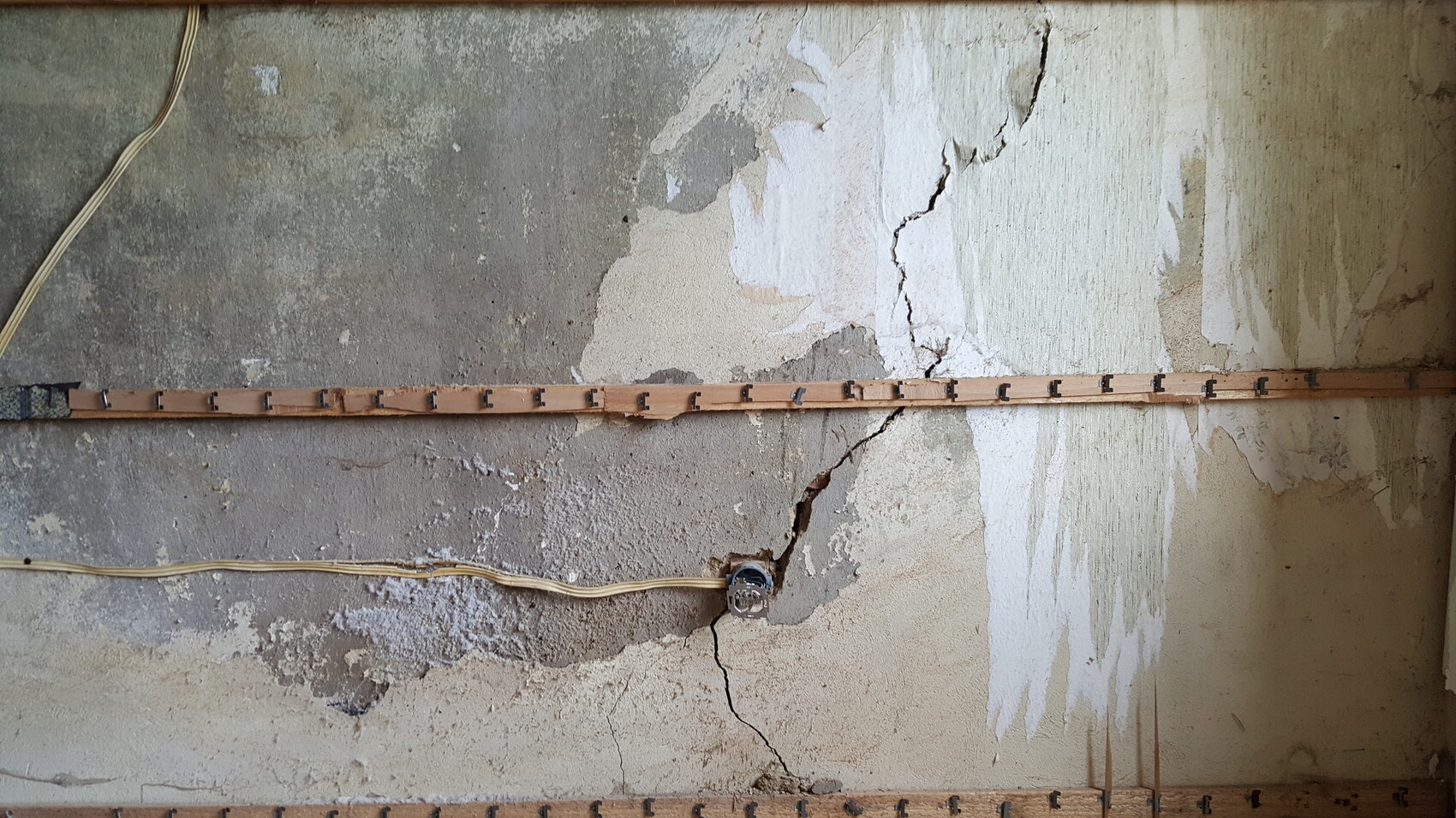 Verwitterte Putzwand mit Rissen und abblätternder Farbe; Holzleiste mit Haken und Kabel.