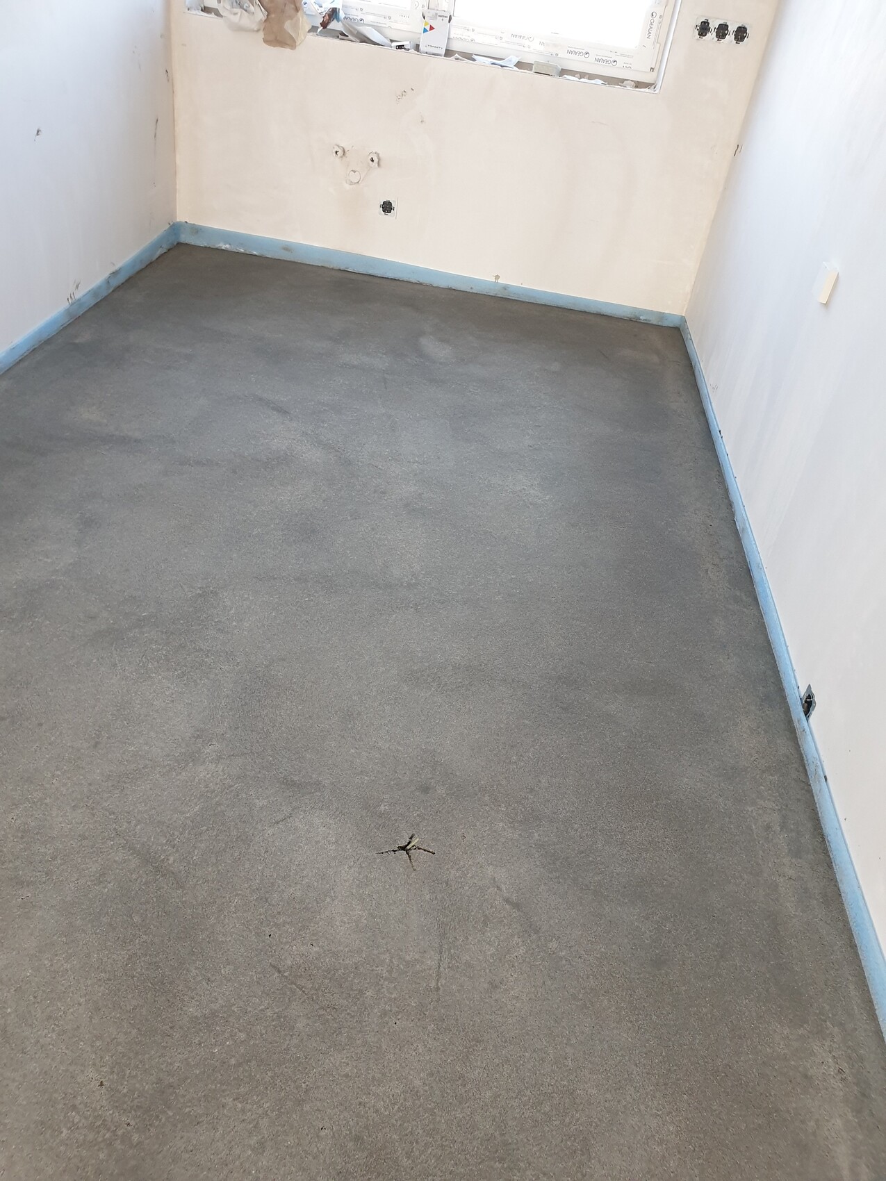 Leerer Raum mit grauem Boden, weißen Wänden, blauer Sockelleiste und offenem Fensterbereich.