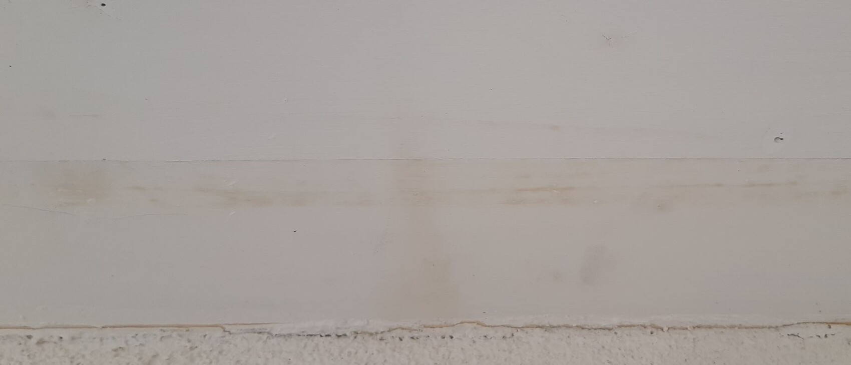 Schlichte hellgraue Wand mit leichten Flecken; unten rauer Boden.