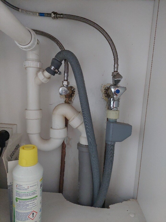 Unter dem Spülbecken: Siphon, Rohre, graue Schläuche und Absperrventil.