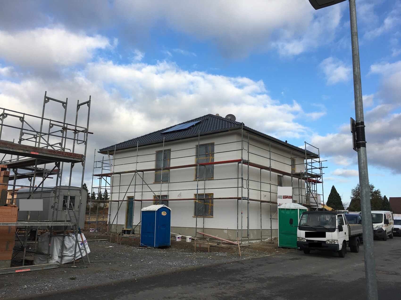 Zweistöckiges Haus im Bau mit Gerüst; Baucontainer, blaue Porta-Potty und parkende Fahrzeuge.