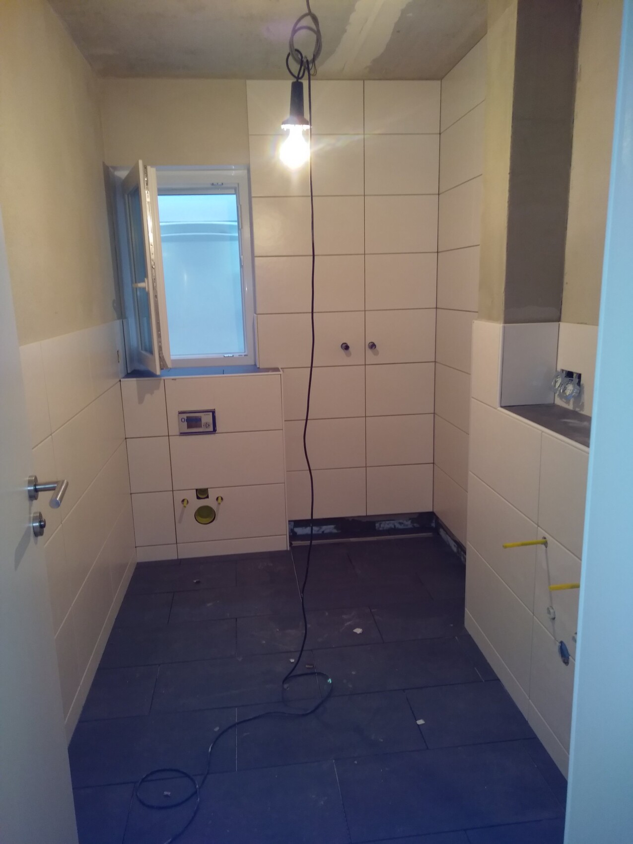 Badezimmer im Bauzustand mit weißen Wandfliesen, offenen Sanitäranschlüssen, Fenster.