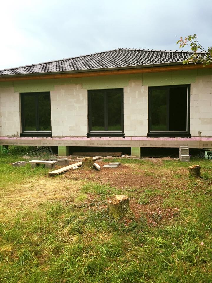 Unvollendete Einfamilienhaus-Baustelle mit drei großen Fenstern, Holzreste und Gras davor.