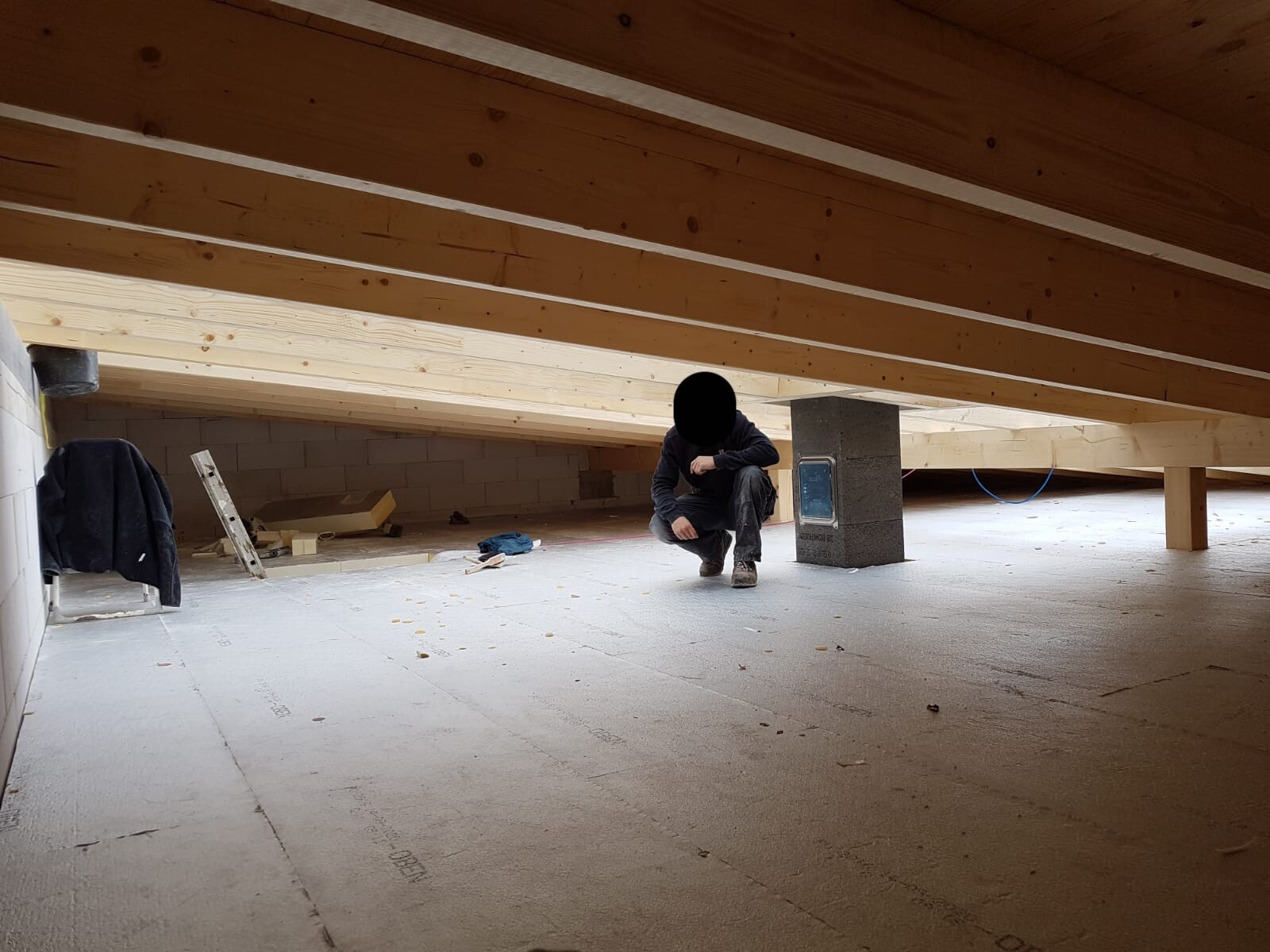Person kniet unter Holzbalkendecke auf einer Baustelle mit Werkzeugen und Dämmplatten
