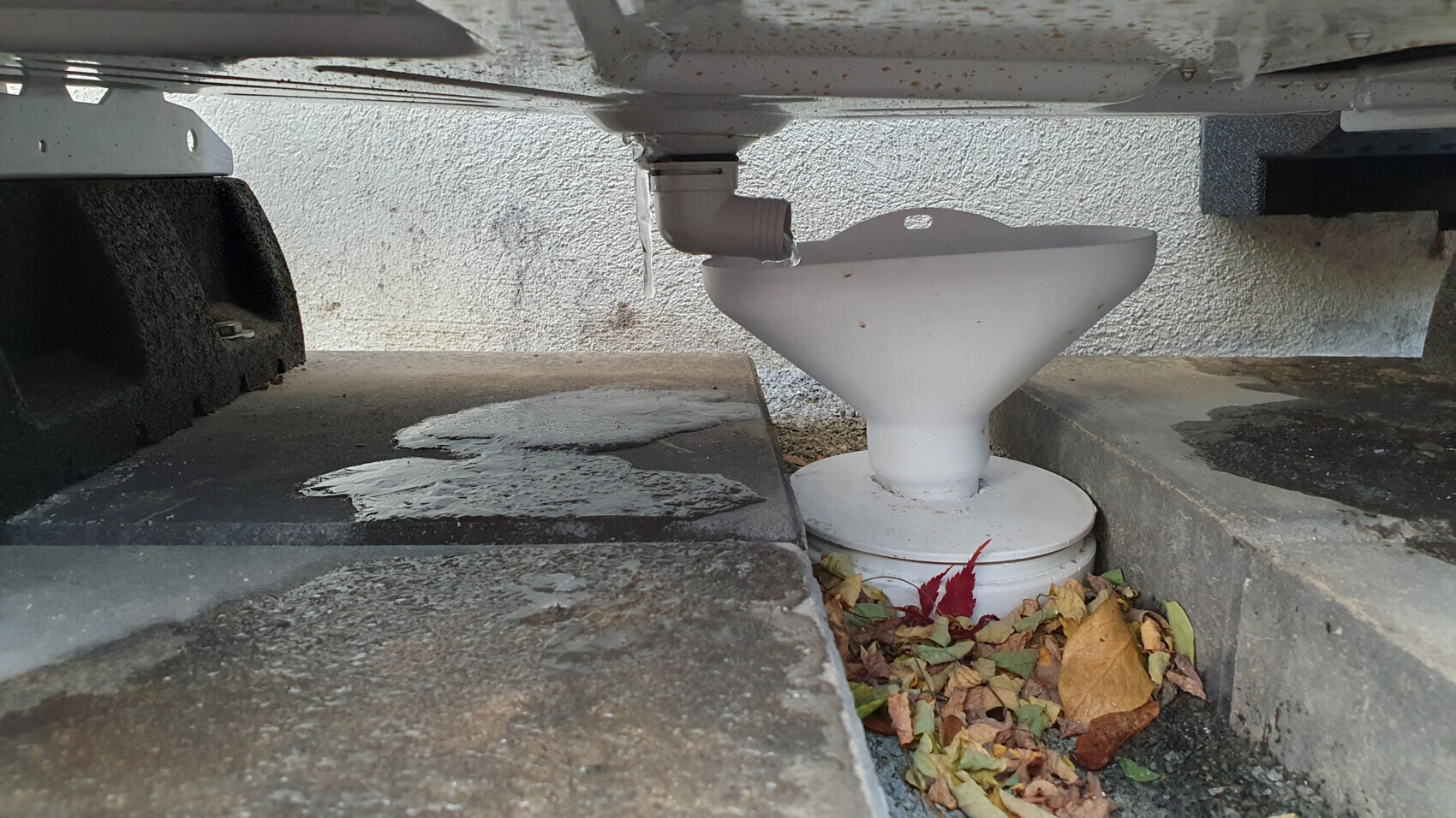 Unter dem Fahrzeug: Weißer Trichter mit Rohranschluss auf Beton, umgeben von Herbstlaub.
