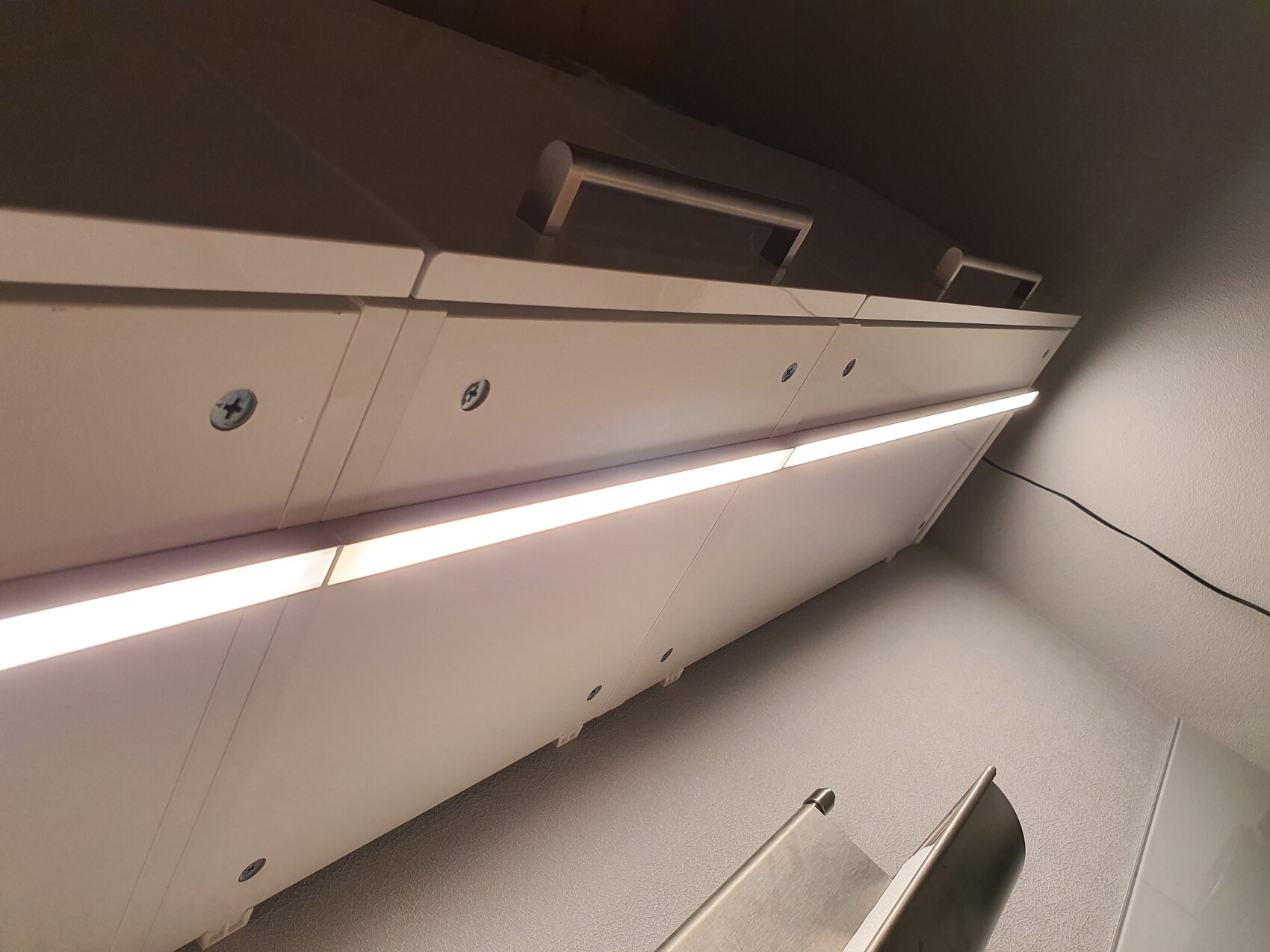 Weiße Schrankfronten mit einer darunter verlaufenden LED-Leiste; Kabel an der Wand.