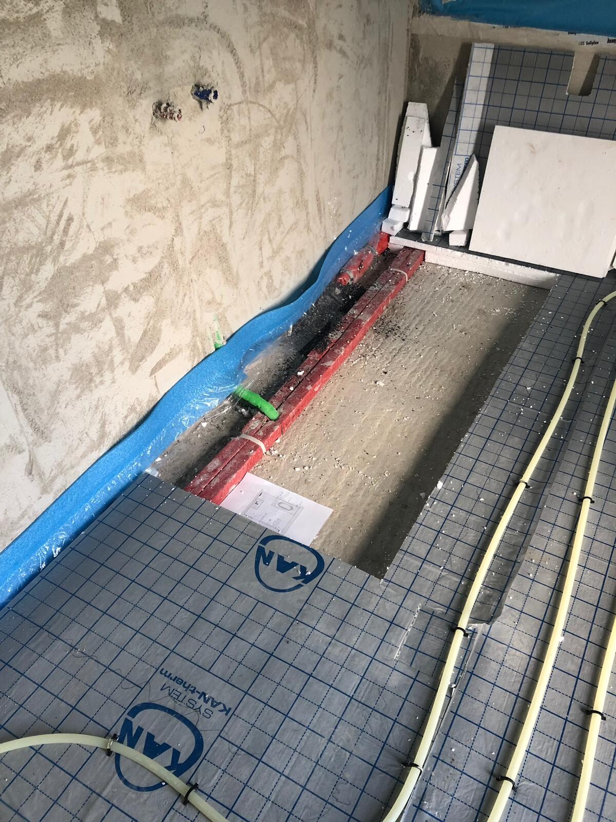 Badezimmer im Bau: blaue Wandabdichtung, gelbe Bodenrohre, Restfliesen und Bauplatten im Raum.