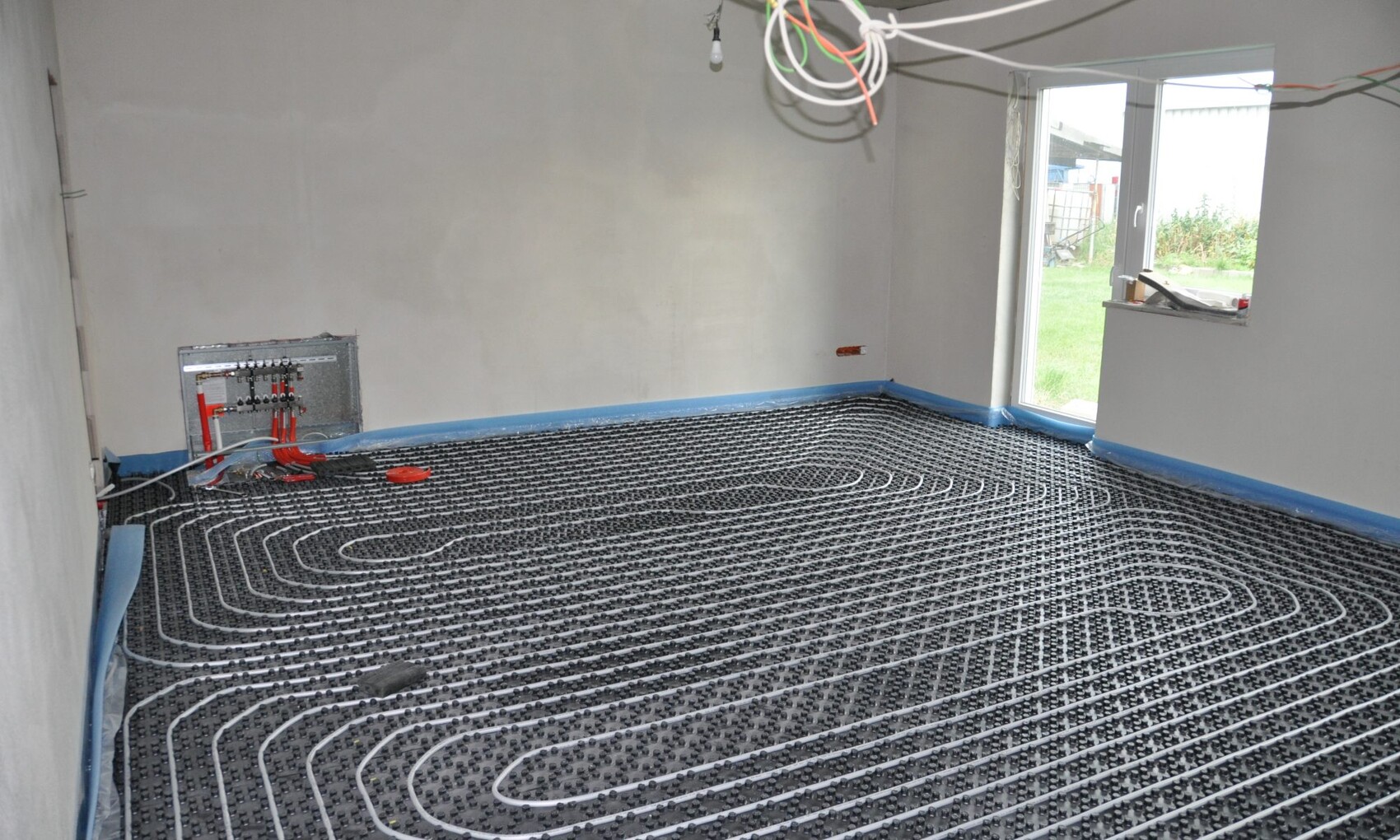 Bauzimmer mit Fußbodenheizung: Heizmatte, Verteiler, Kabel, offenes Fenster.