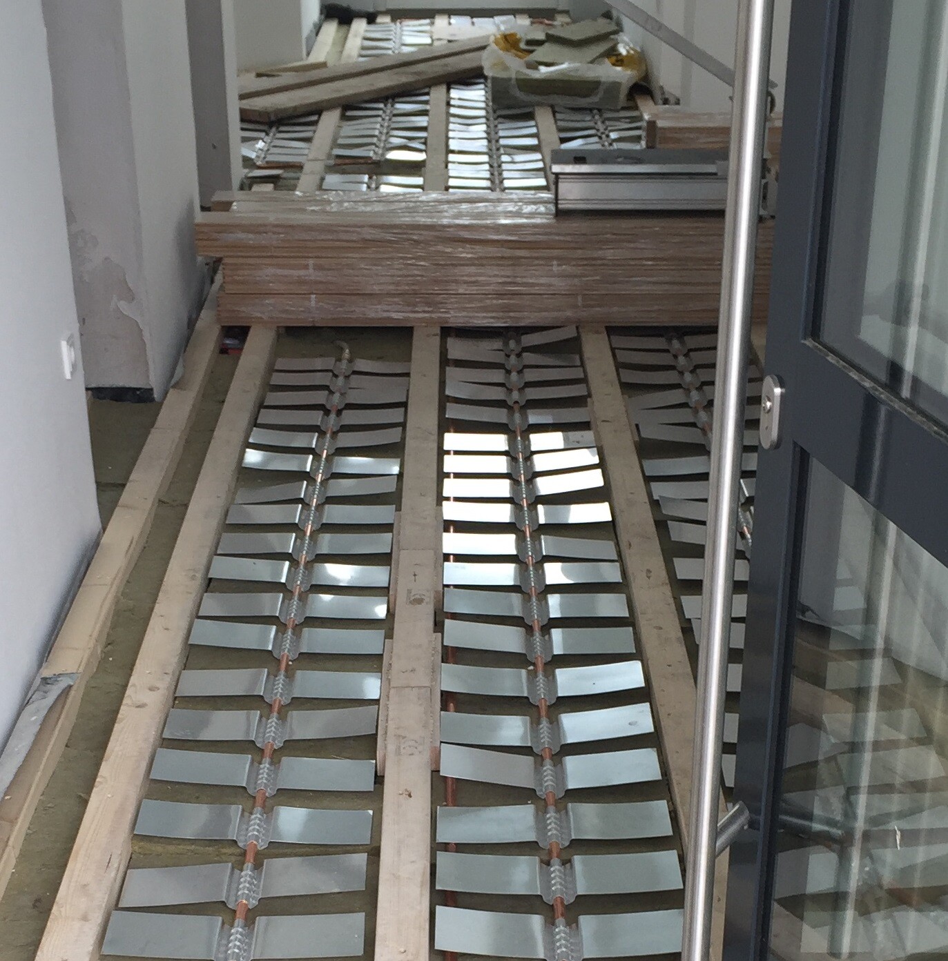 Innenraum einer Baustelle: Holzlatten liegen auf dem Boden, Metall-Lamellen in Reihen, Tür rechts.