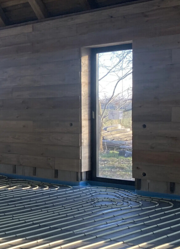 Innenraum im Bau: Fußbodenheizungsrohre, Holzverkleidung, schmale Tür mit Blick nach außen.