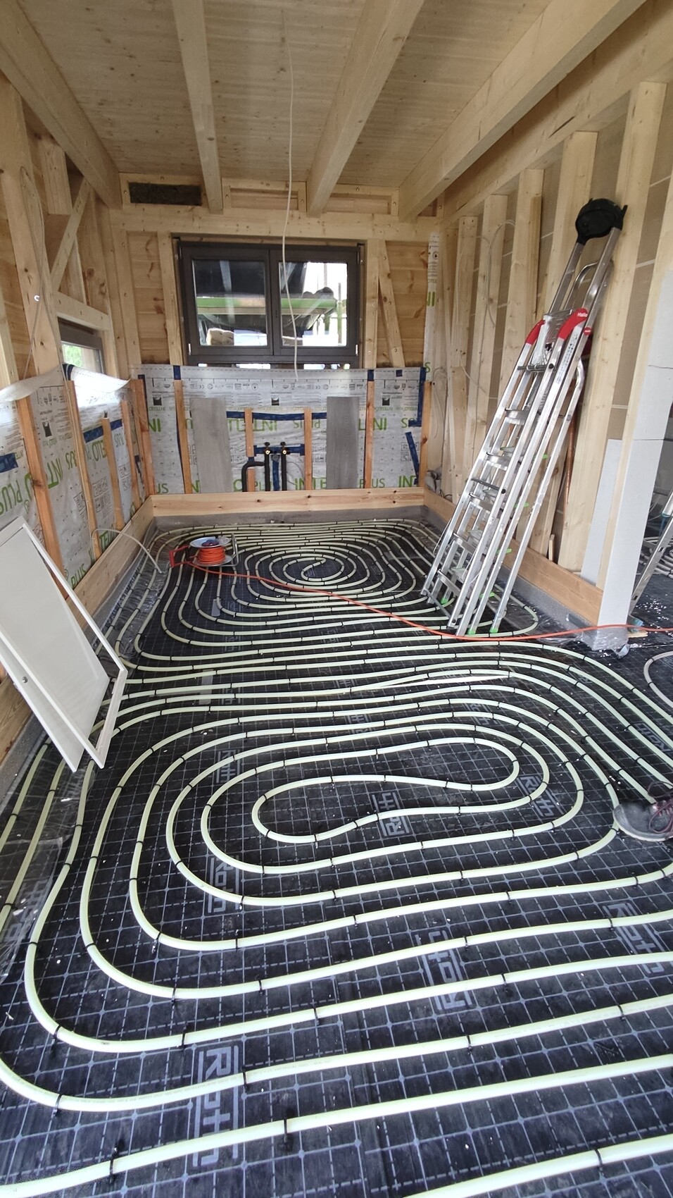 Rohbauzimmer mit sichtbarer Fußbodenheizung: spiralförmig verlegte Rohre im Boden, Holzrahmen.