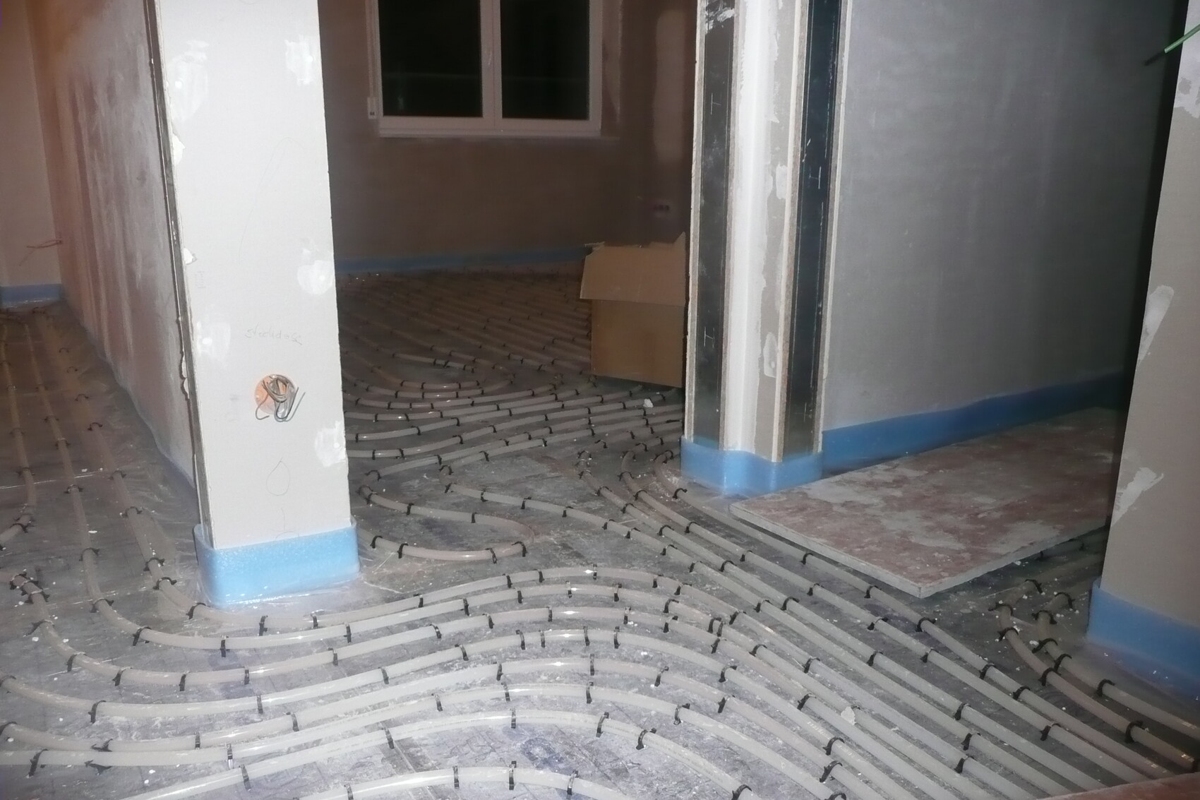 Unterbodenheizung: Rohrschlangen im Rohbau, blaue Sockelleiste, Fenster im Hintergrund.