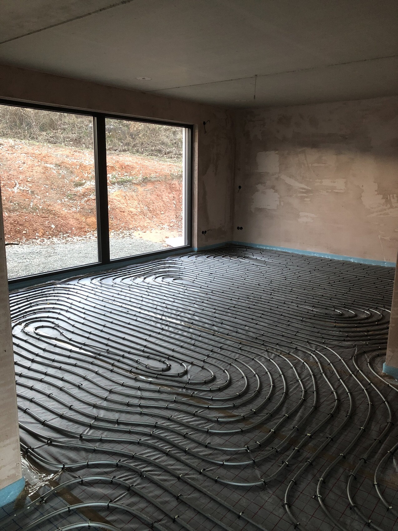 Rohbauzimmer mit spiralförmig verlegten Fußbodenheizungsrohren und großen Fenstern.