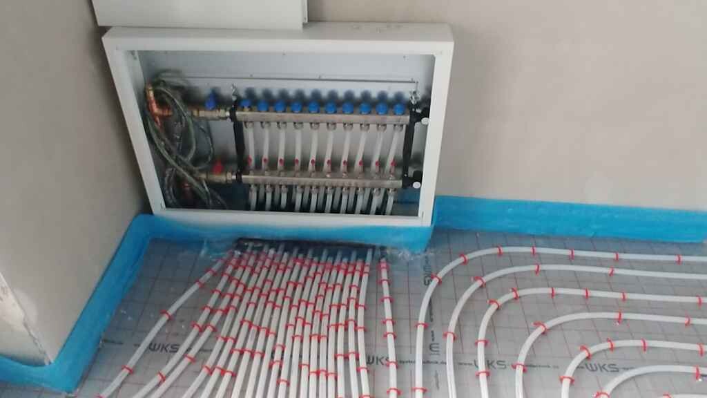 Unterflur-Heizungsrohre spiralförmig auf dem Boden, daneben Wand-Verteilerkasten mit Ventilen.