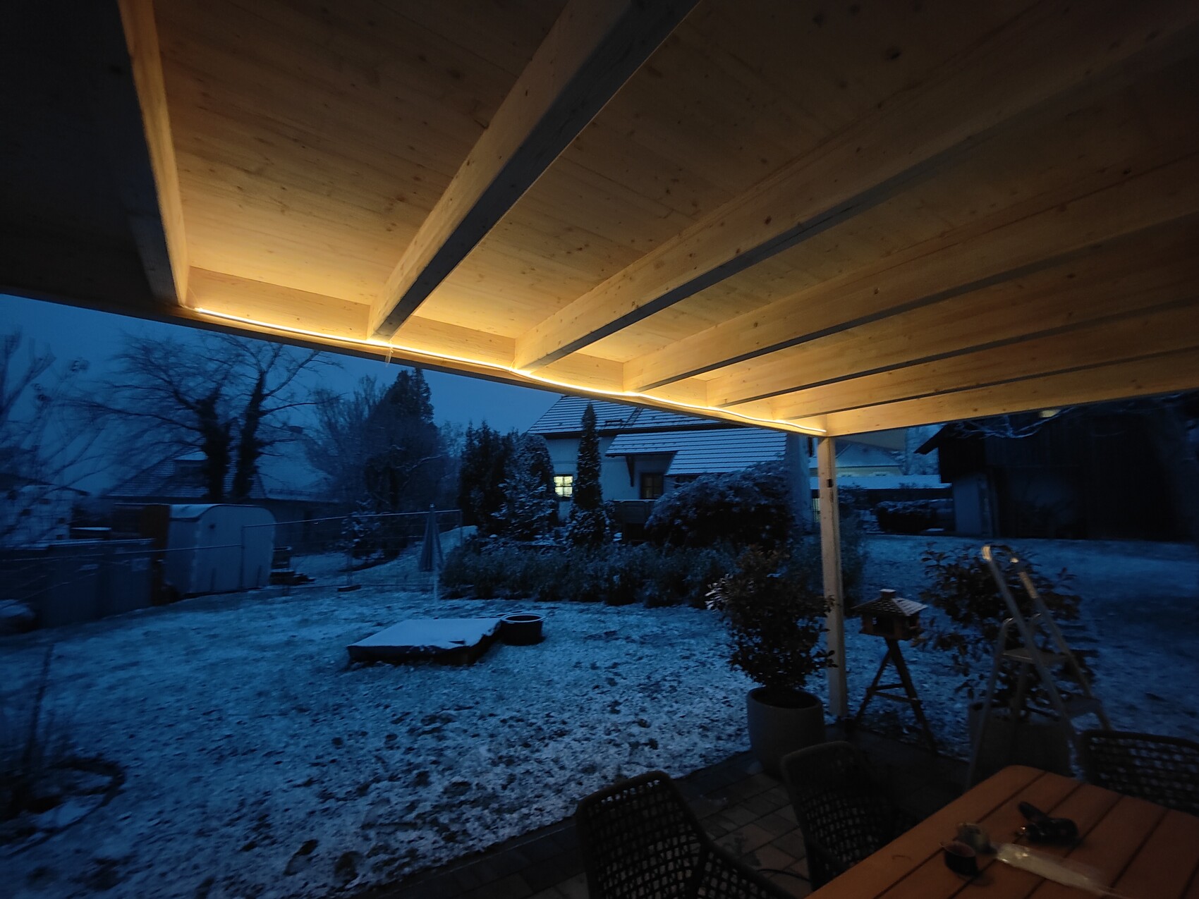 Überdachte Holzveranda bei Dämmerung; warmes Gelblicht, schneebedeckter Garten mit Möbeln.