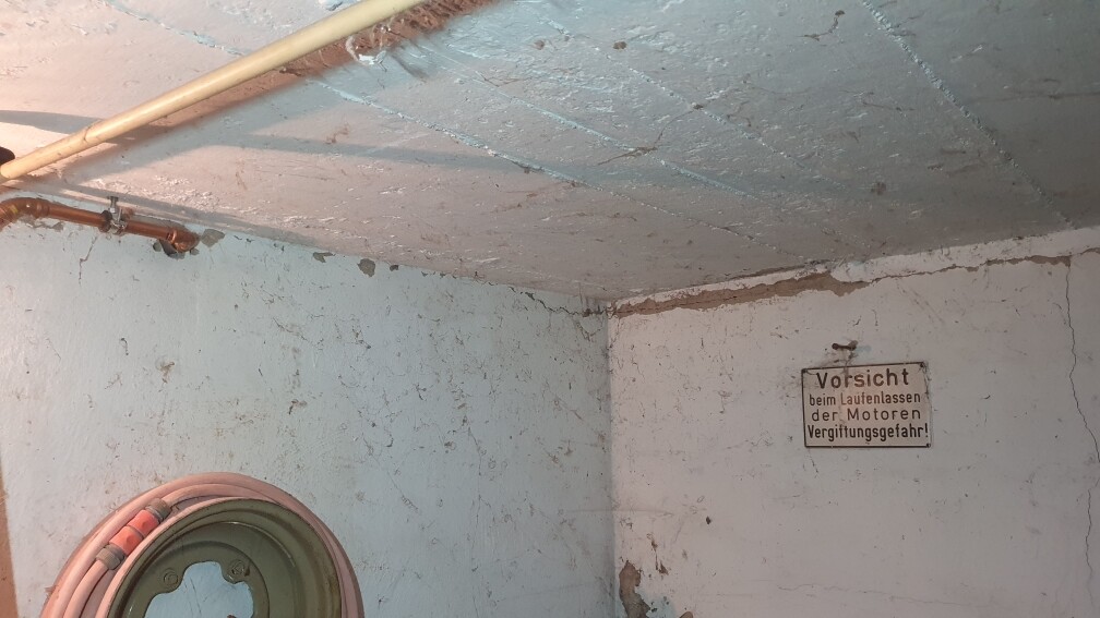 Kellerraum mit Rohren an der Decke, aufgewickelter Schlauchrolle und Warnschild vor Vergiftungsgefahr.