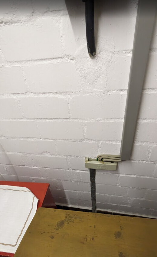 Weiße Ziegelwand mit senkrechtem Kabelkanal, Klemmkasten und Holzunterlage im Vordergrund.