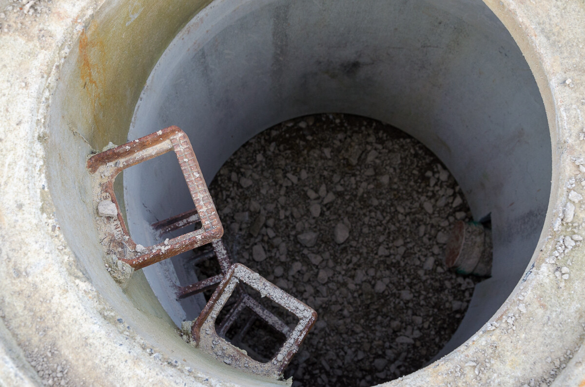 Rostige Metallleitern im runden Betonschacht, Boden mit Kies und Steinen.
