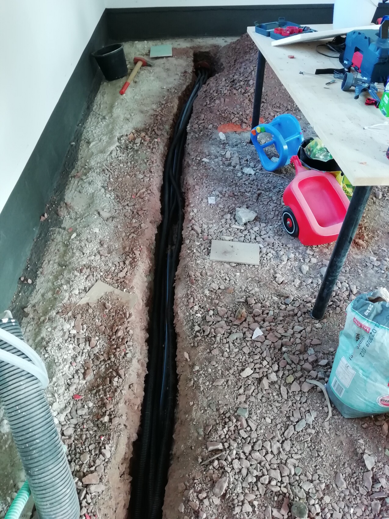 Baugrube neben Wand mit schwarzen Kabeln; Spielzeugauto und Lenkrad-Spielzeug liegen daneben.