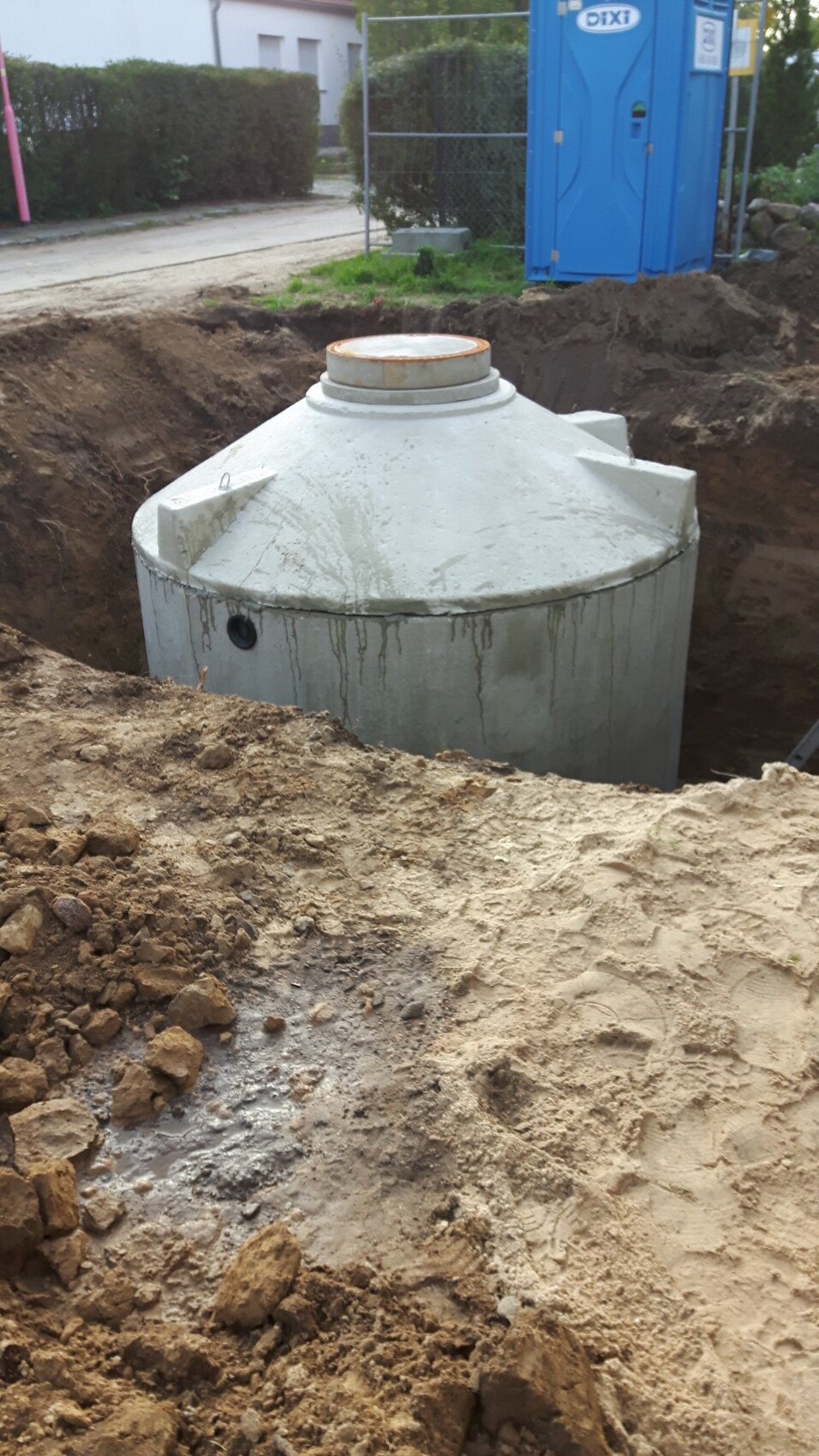 Großer grauer Betontank in Baugrube, Baustelle; daneben blaue Mobiltoilette.