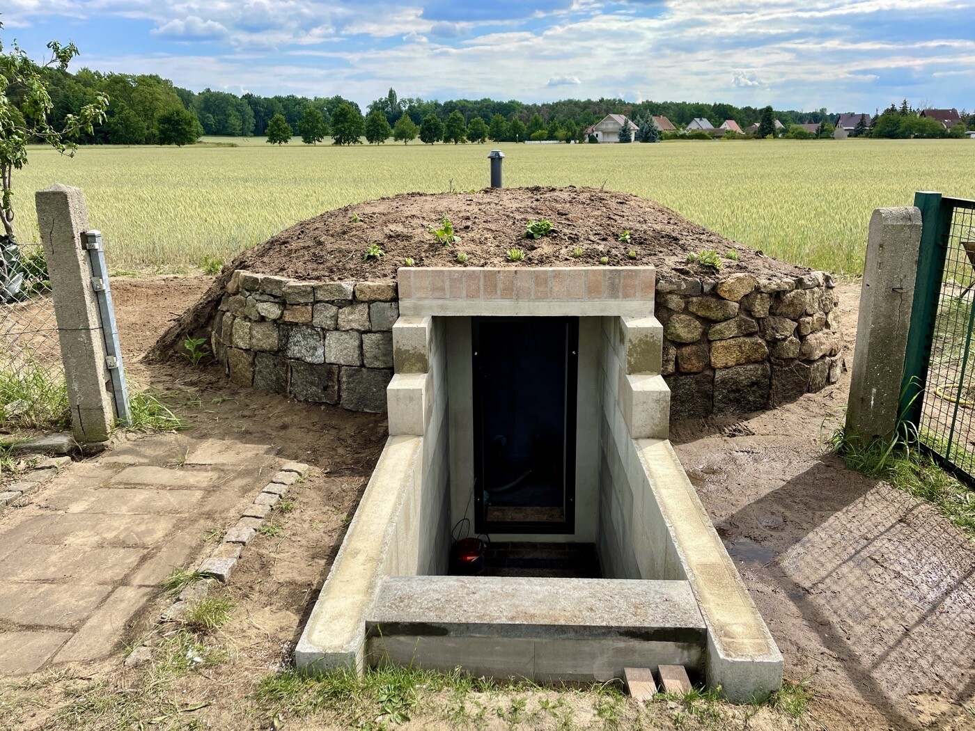 Unterirdischer Kellerzugang mit Steinmauer, Erdhügel und offener Eingang im Feld