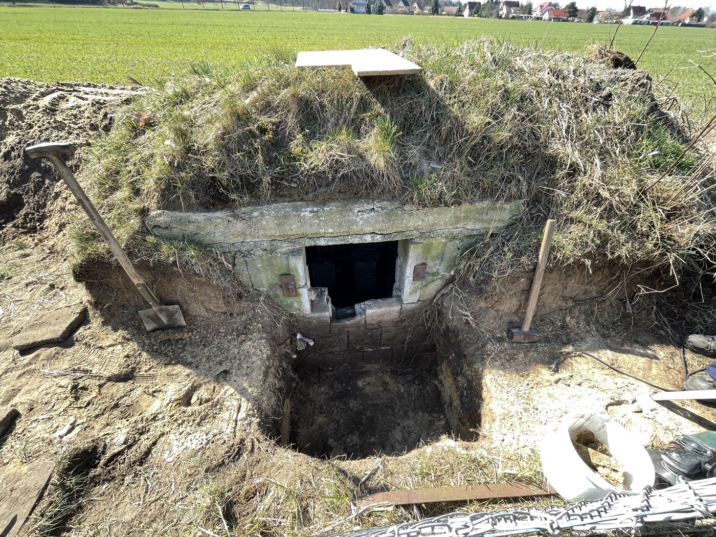 Grubenöffnung im Erdwall mit Betonrahmen; dunkler Schacht, Gras darüber, ländliche Felder dahinter.