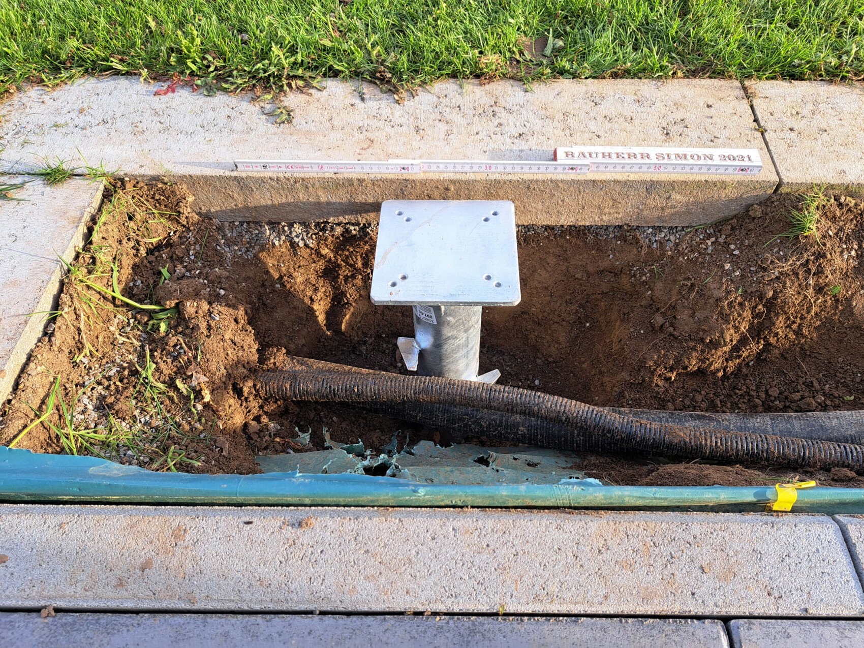 Offene Baugrube neben Gehweg mit Metallpfosten und quadratischer Platte; Kabelschlauch liegt darin.