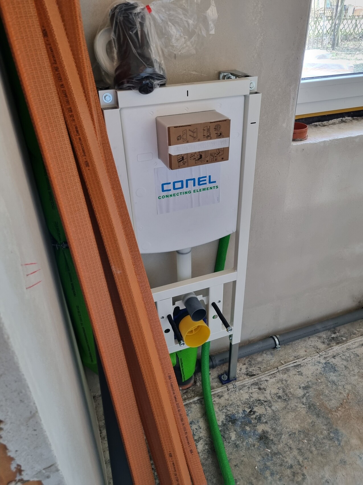 Weißer Wand-WC-Rahmen mit Karton-Verpackung; orange Bauplatten und grüne Schläuche daneben.