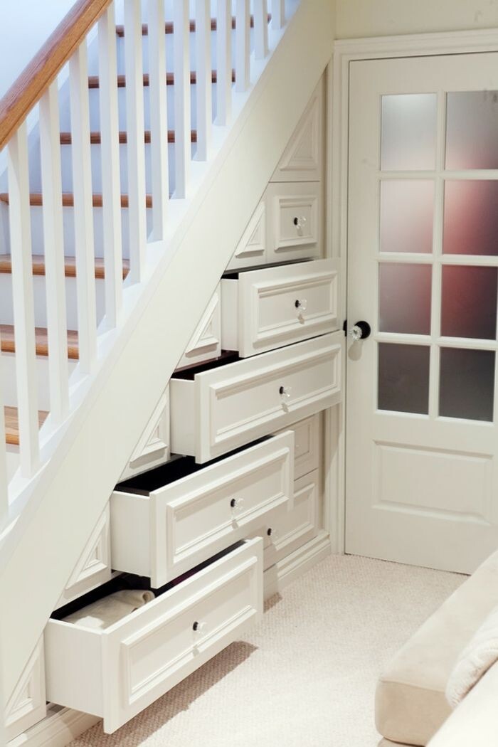 Unter der Treppe befinden sich mehrere weiße Schubladen, daneben eine Glastür.