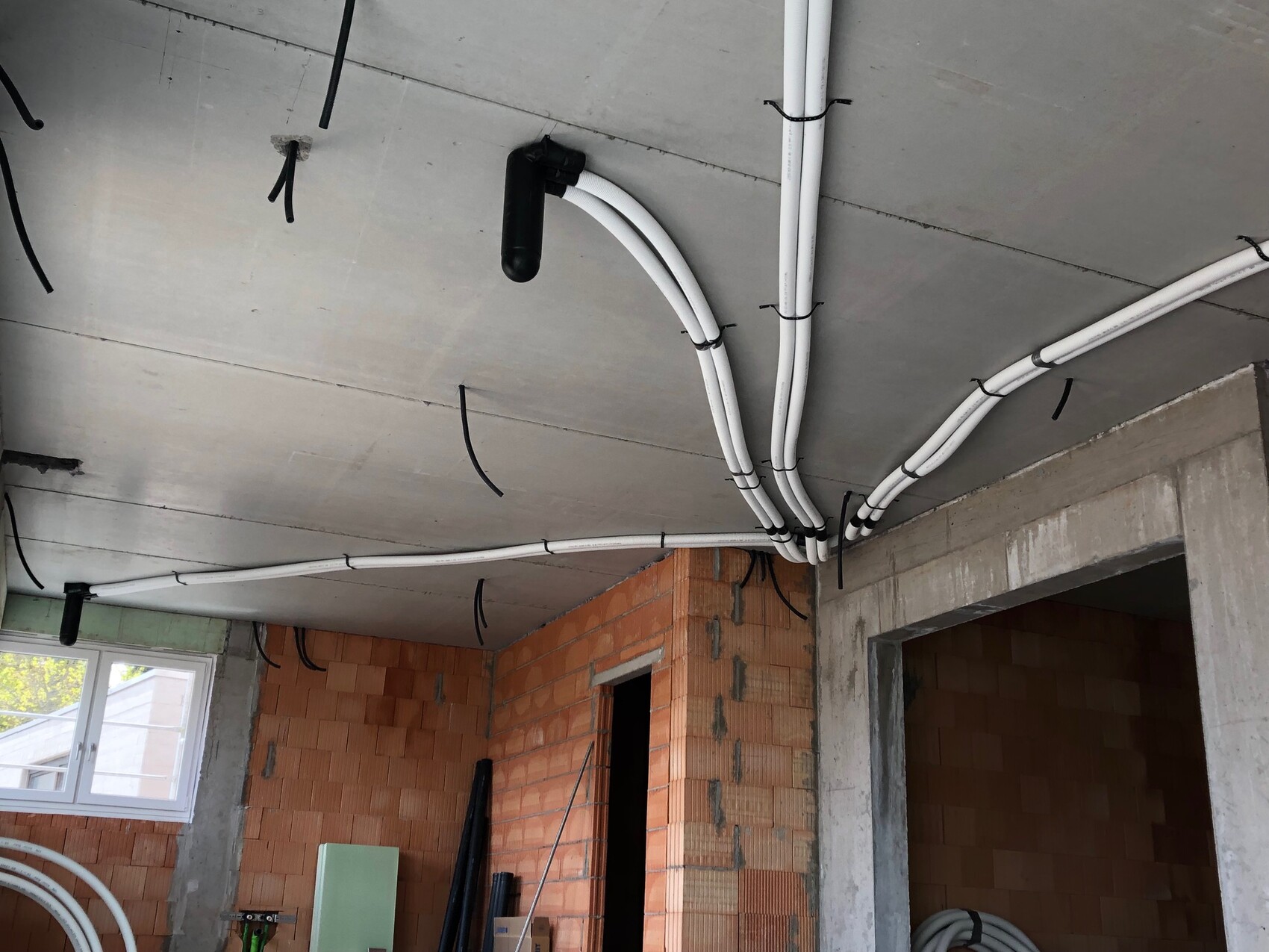 Unfertiger Raum: Weiße Kabelschläuche verlaufen an der Decke, schwarze Halterungen, Ziegelwände.