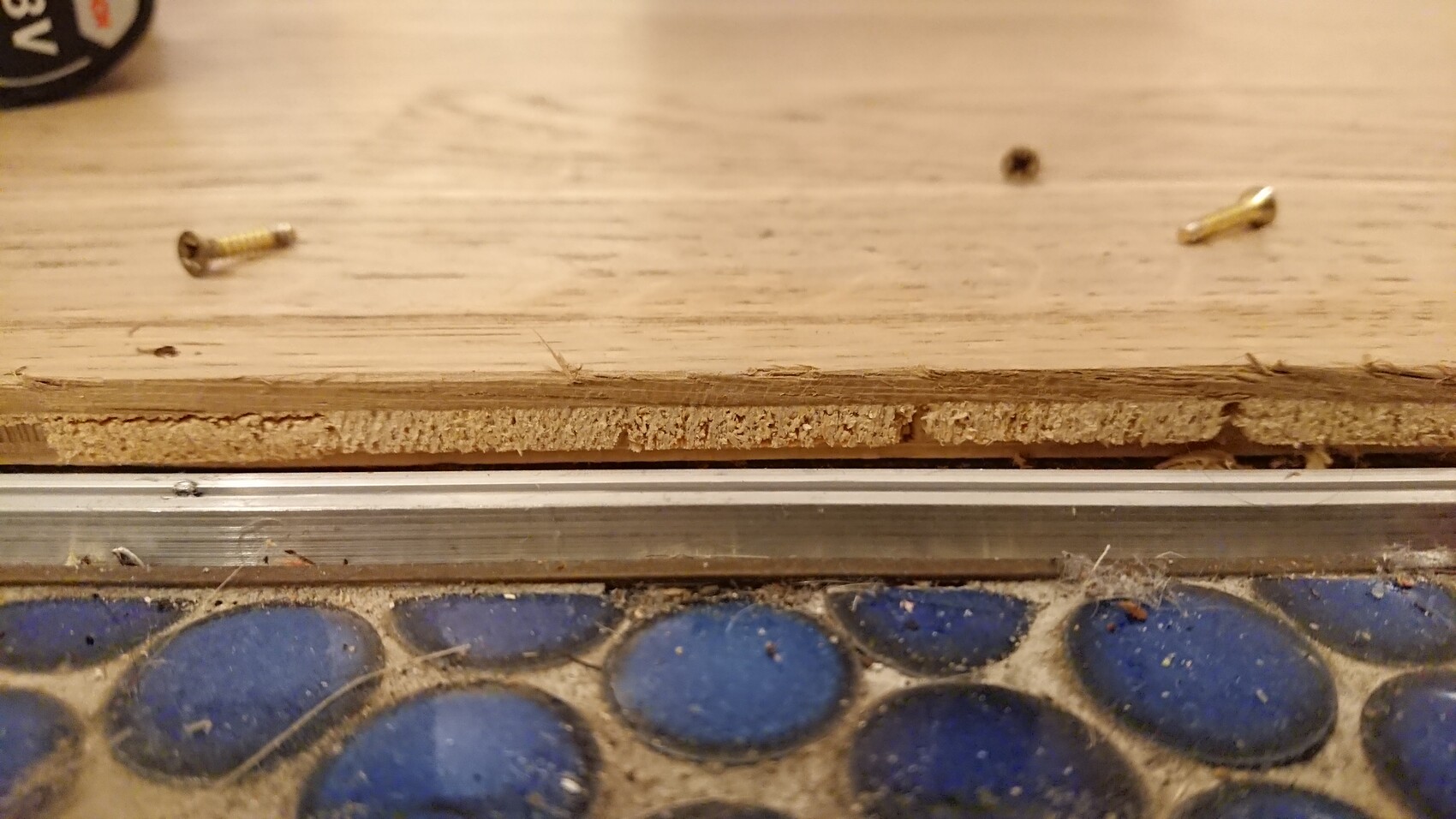 Nahaufnahme einer Holzplattenkante mit Spanplattenkern; zwei Schrauben liegen auf dem Boden.