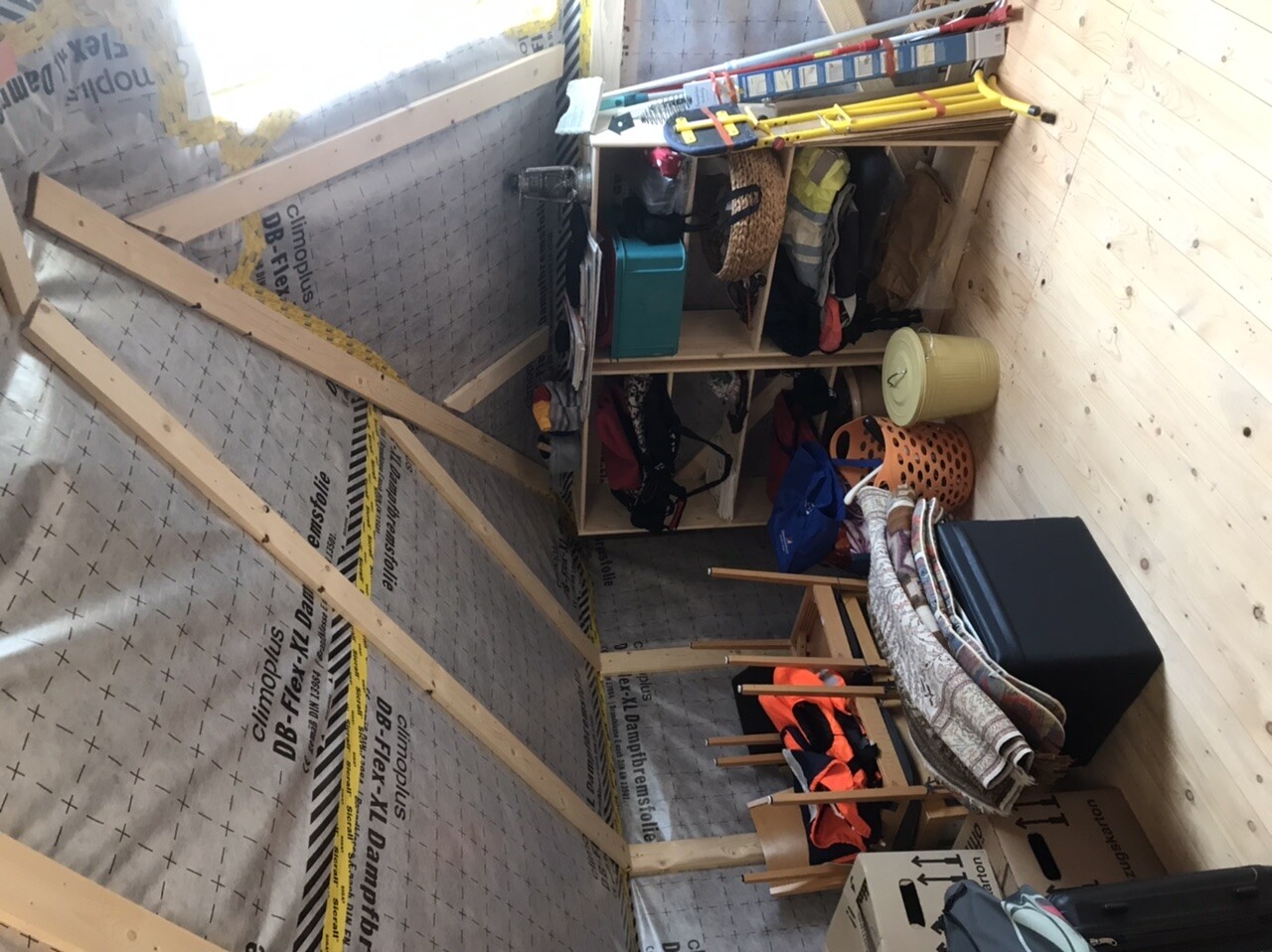Unfertiger Abstellraum mit Regal, Kleidung, Körben, Werkzeugen und Dämmplatten an der Wand.