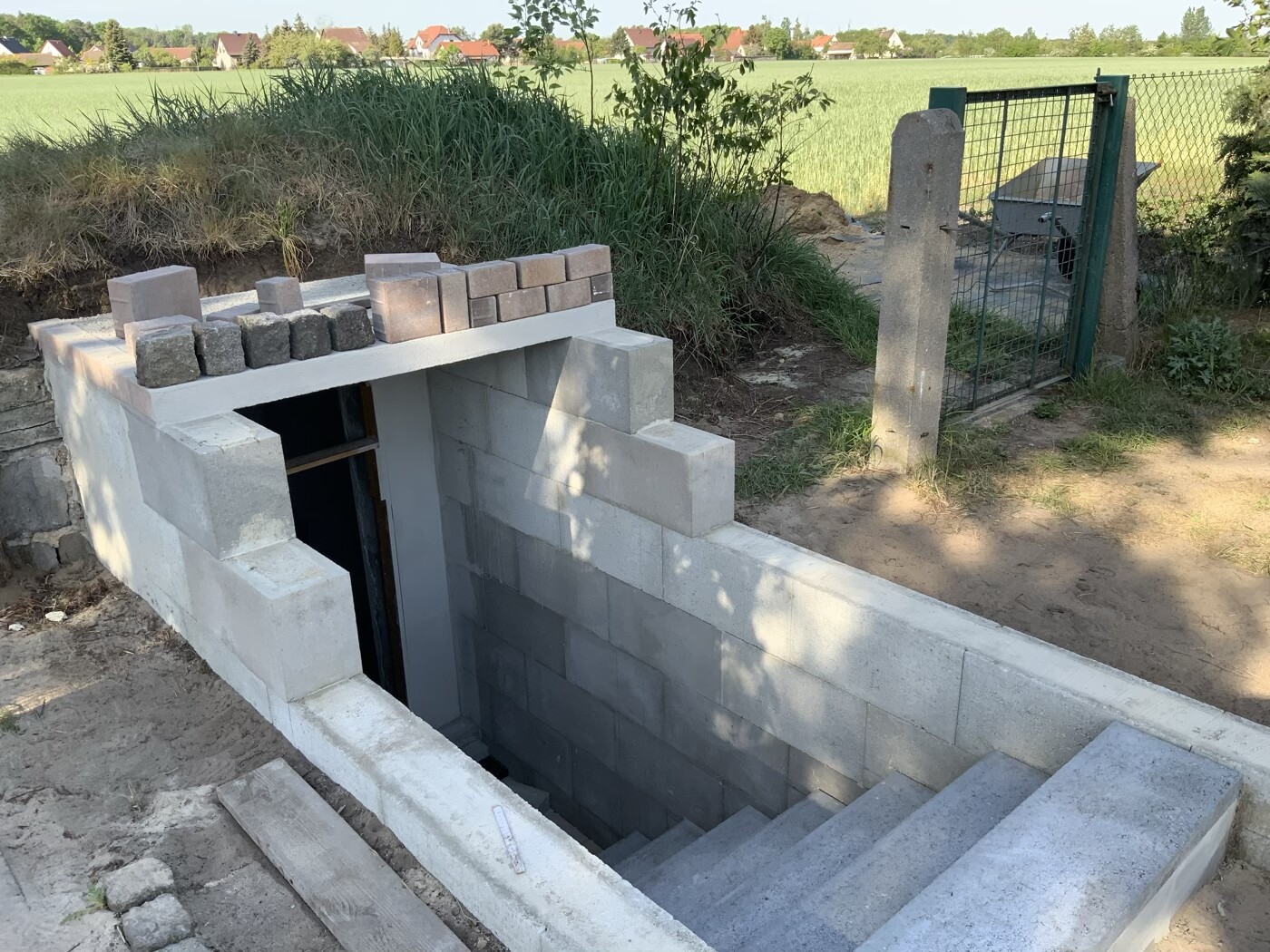 Beton-Treppenabgang in Erdwall; Blöcke oben gestapelt, Zaun und Felder im Hintergrund.