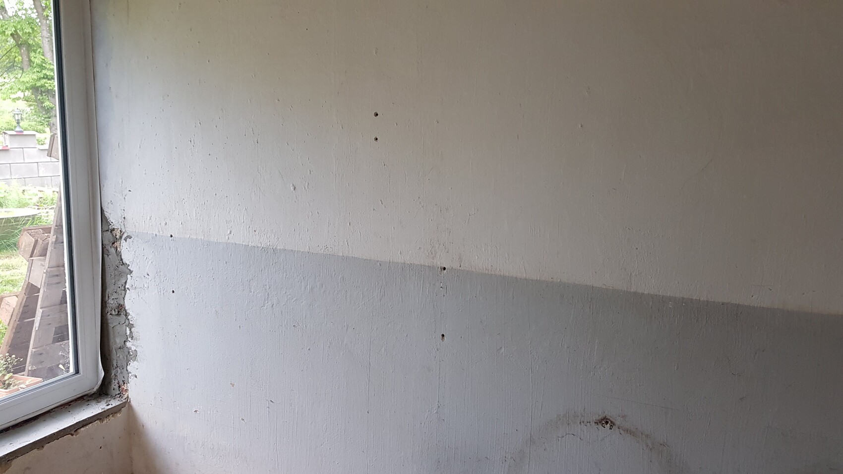 Helle Oberseite und graue Unterseite einer Wand mit Löchern; links Fenster mit Außenblick.