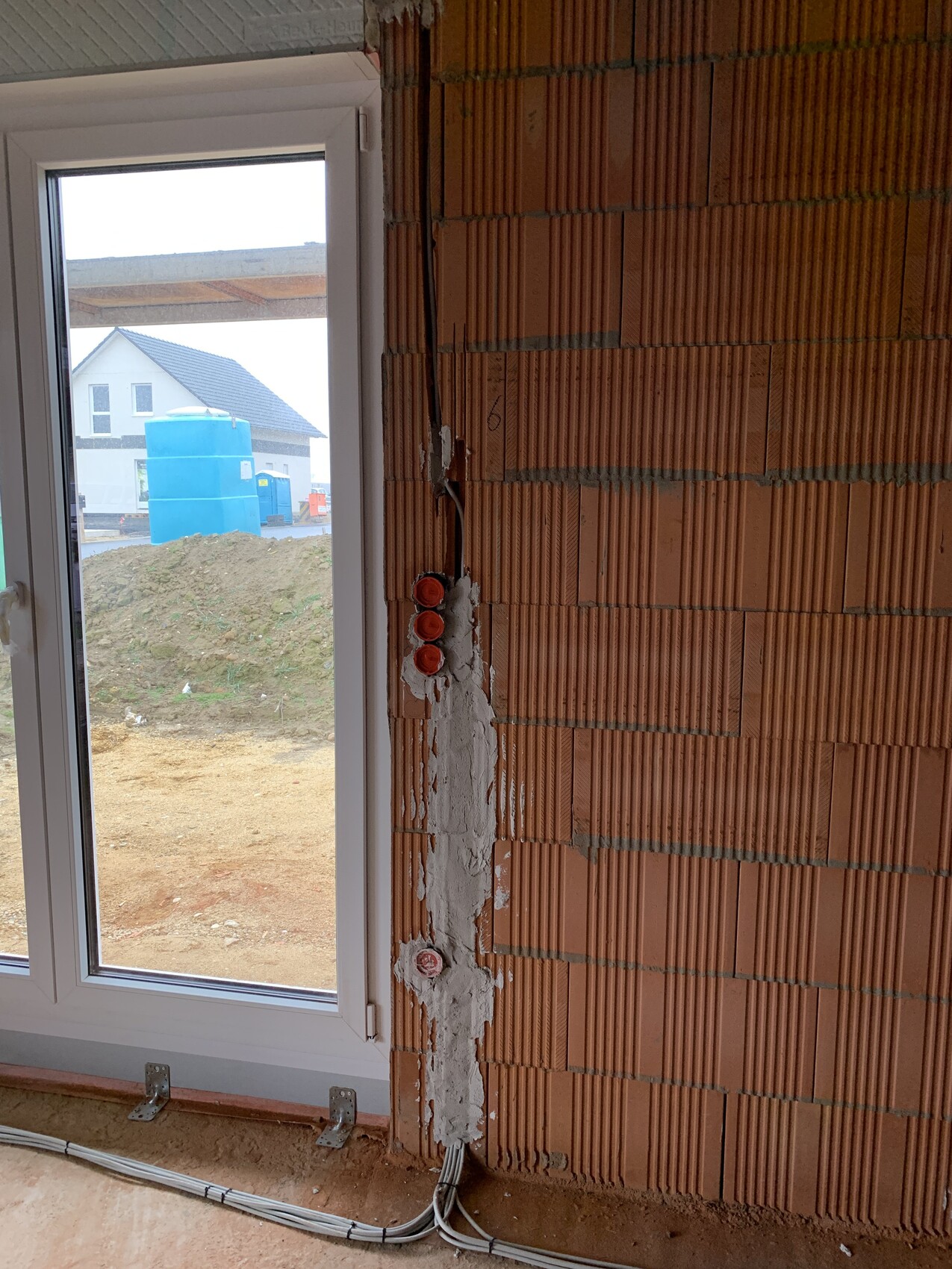 Rohbau-Innenraum: Ziegelwand, freiliegende Kabel; Fenster zeigt Außenansicht mit blauem Wassertank.