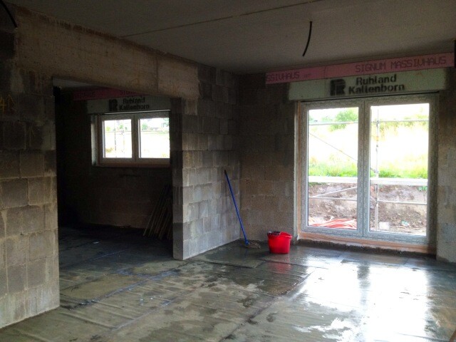 Unfertiger Bauinnenraum: Betonmauern, großes Fenster, Besen, roter Eimer, feuchter Boden.