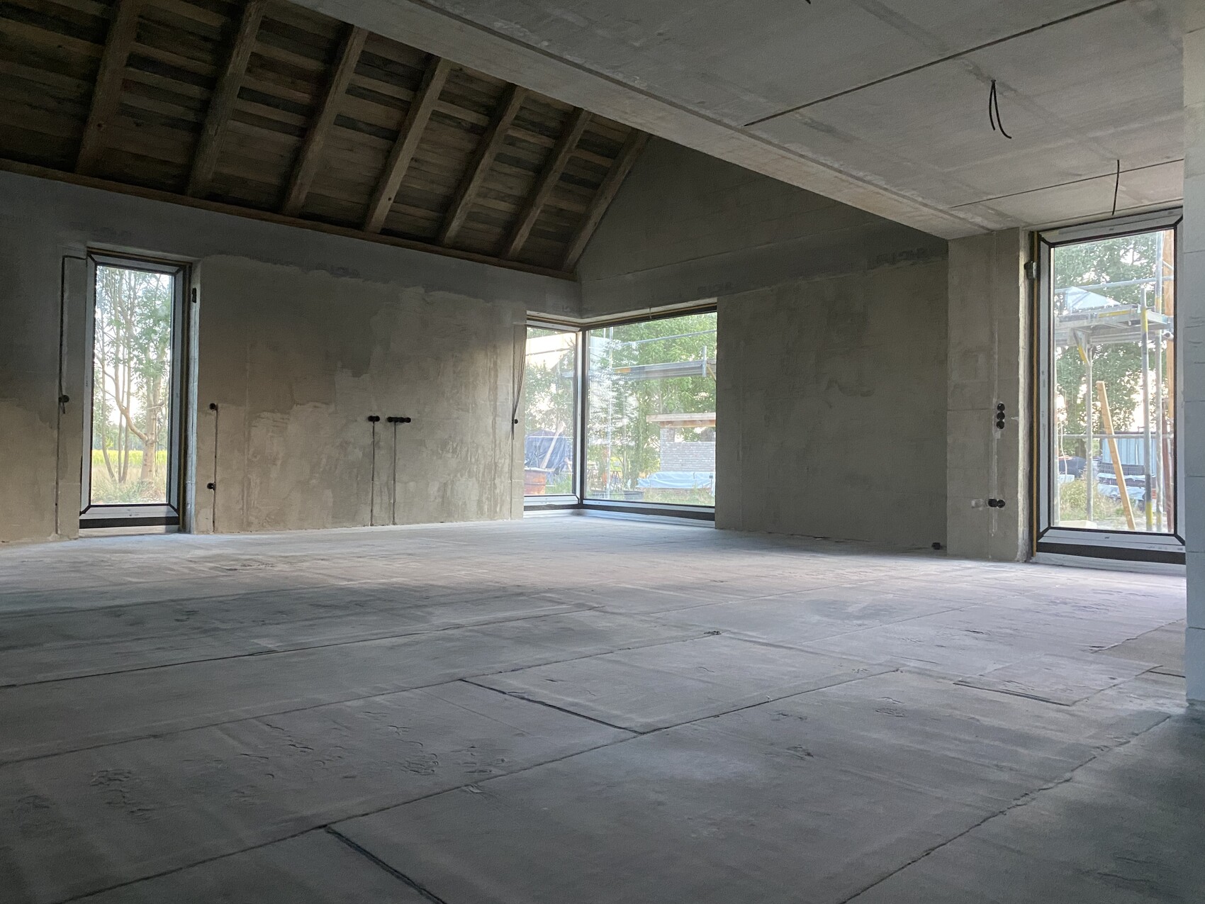 Offener Rohbau-Raum mit Betonboden, rauen Wänden, sichtbaren Dachbalken und großen Fenstern.