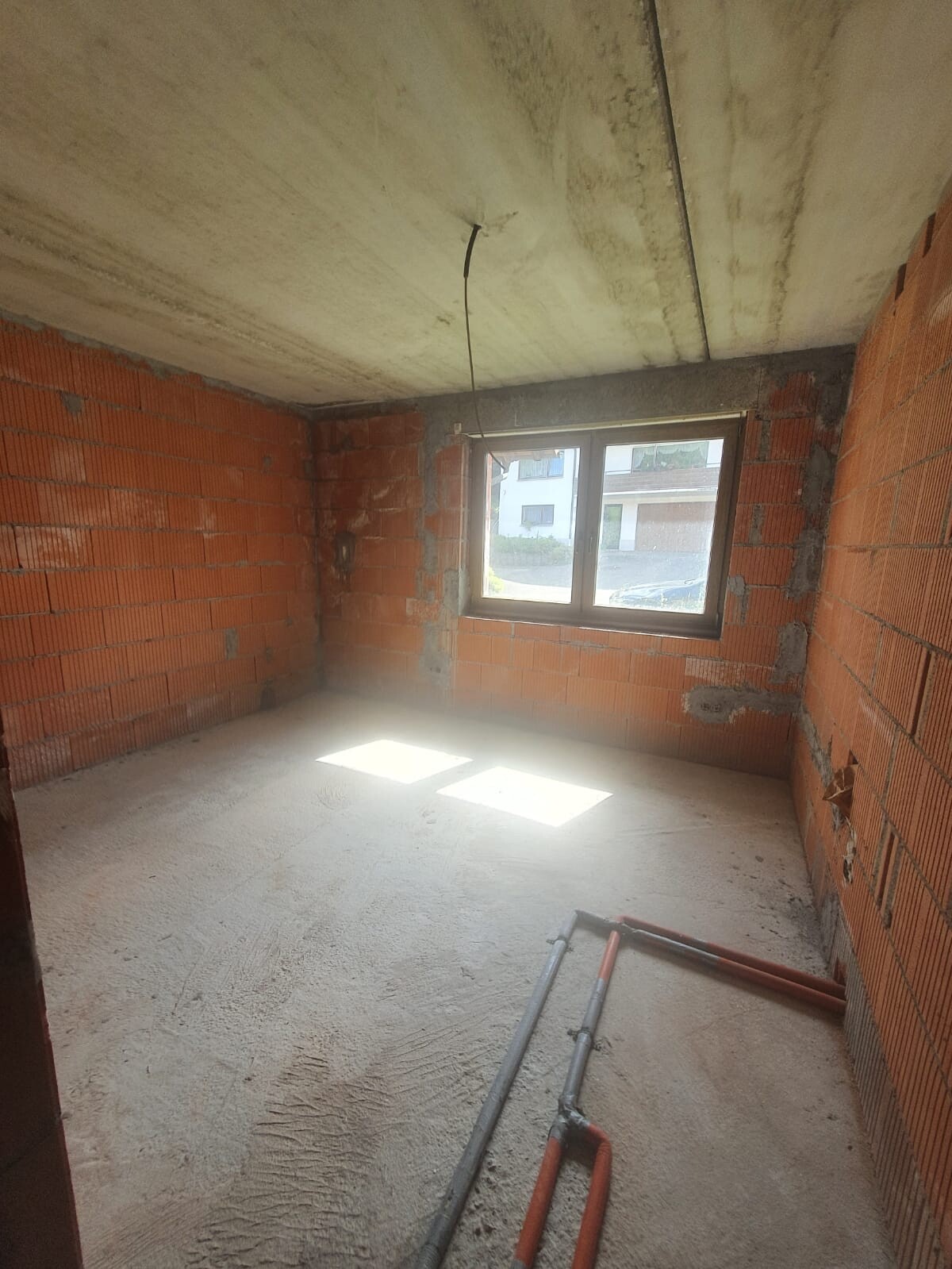 Unverputzter Rohbau-Raum mit Ziegelwänden, Beton-Decke, Fenster und Rohren am Boden.